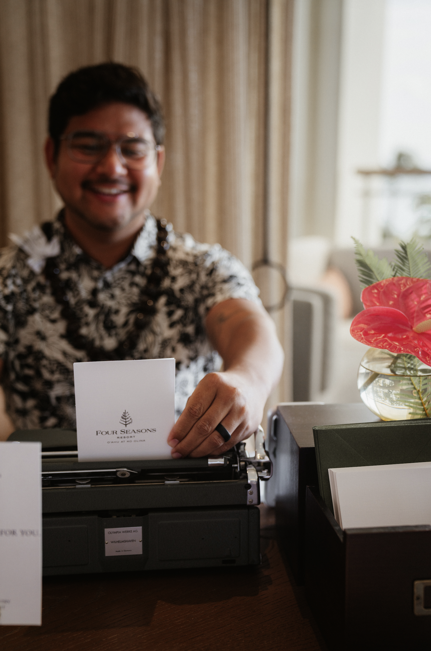 A smiling man wearing glasses and a black and white patterned shirt sitting at a desk with a typewriter, displaying a card that reads "Four Seasons Resort". There is a vase with red and green flowers on the right side of the image, and the background shows beige curtains and a window with bright natural light.