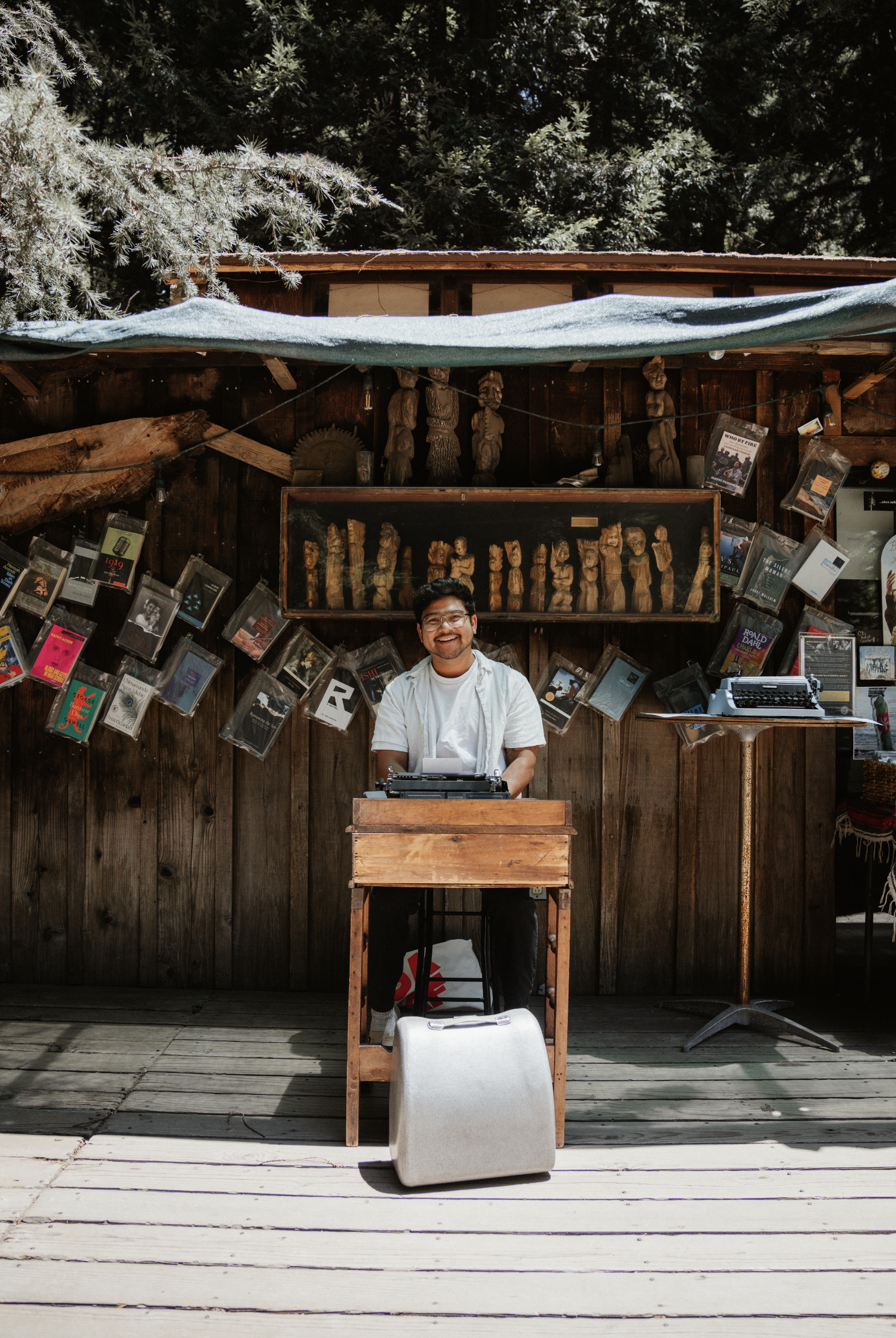 A man standing behind a wooden desk or table outdoors, possibly at a market or event, with books and wooden carvings displayed behind him. He is smiling, wearing glasses and a white shirt, and has a typewriter on the table. There is a large, light-colored suitcase or bag in front of him on a wooden deck, with trees in the background.