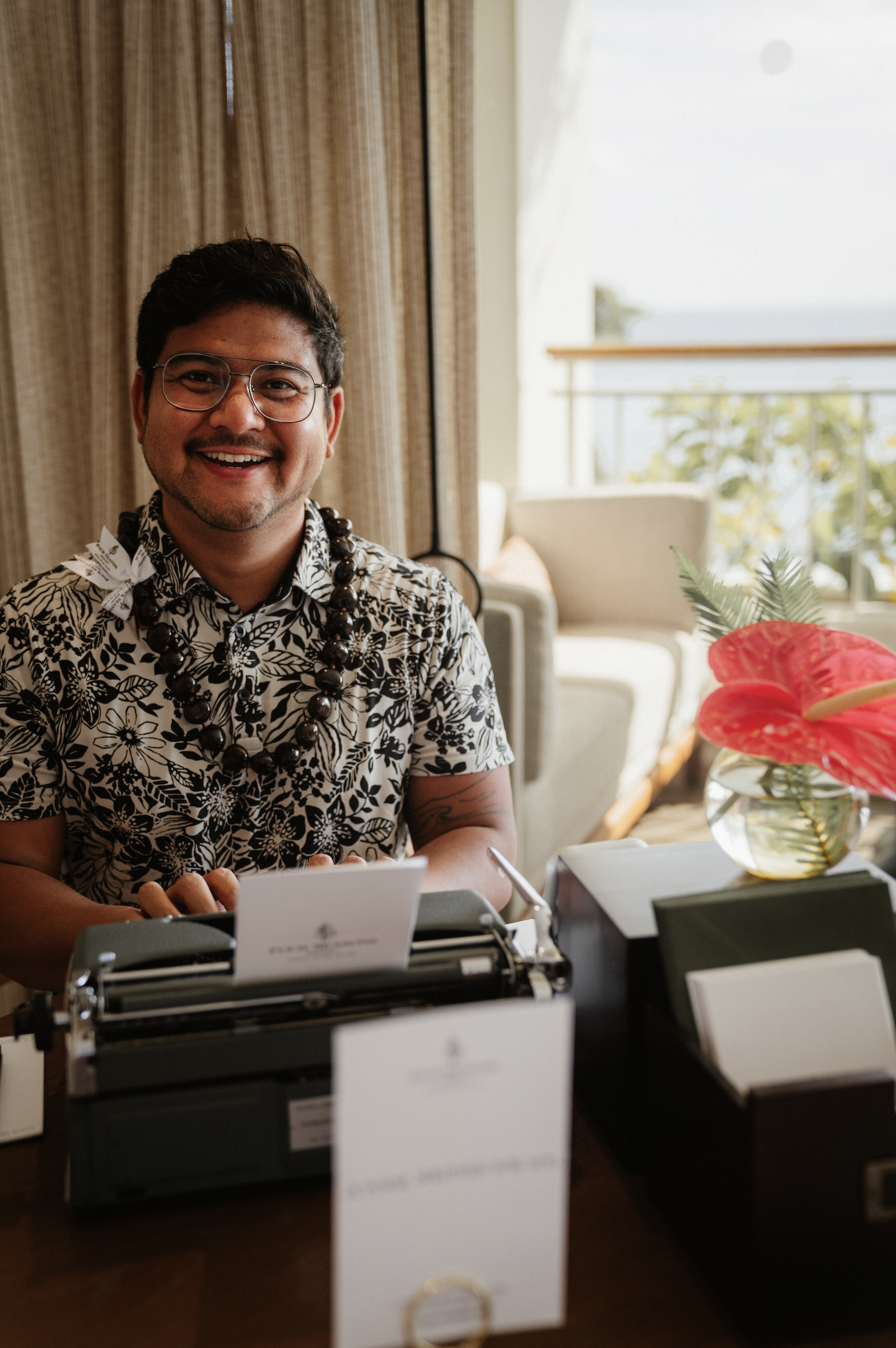 A man with dark hair, glasses, and a smile, wearing a black and white floral shirt and a beaded necklace, is sitting at a table with a typewriter, various papers, and a potted plant with red leaves in a glass vase. The background features curtains, a window with a view of trees, and a beige armchair.