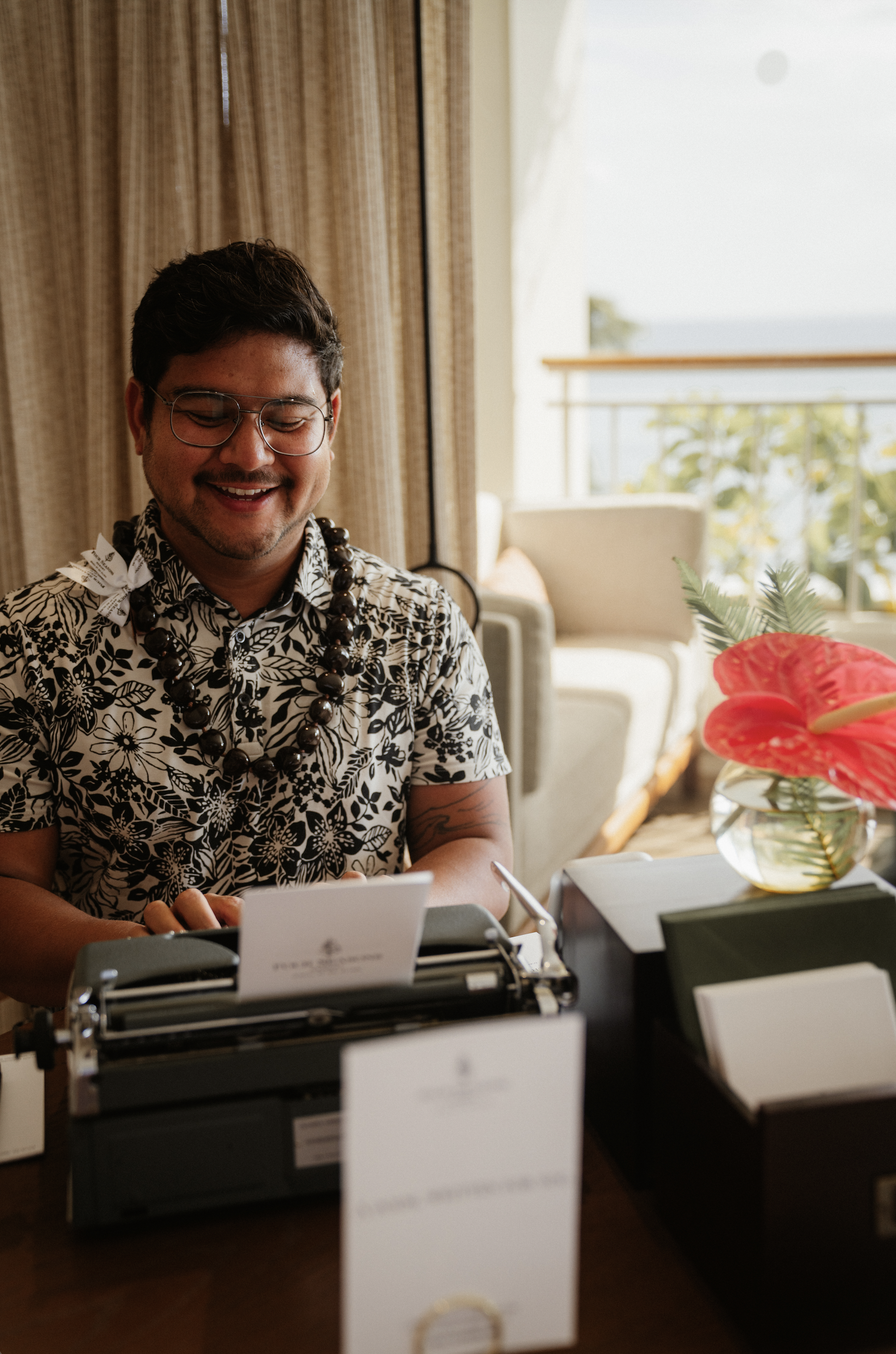 A man wearing glasses and a floral shirt, smiling, sitting at a desk with a vintage typewriter, surrounded by papers and a vase with tropical flowers, in a well-lit room with window and beige curtains.