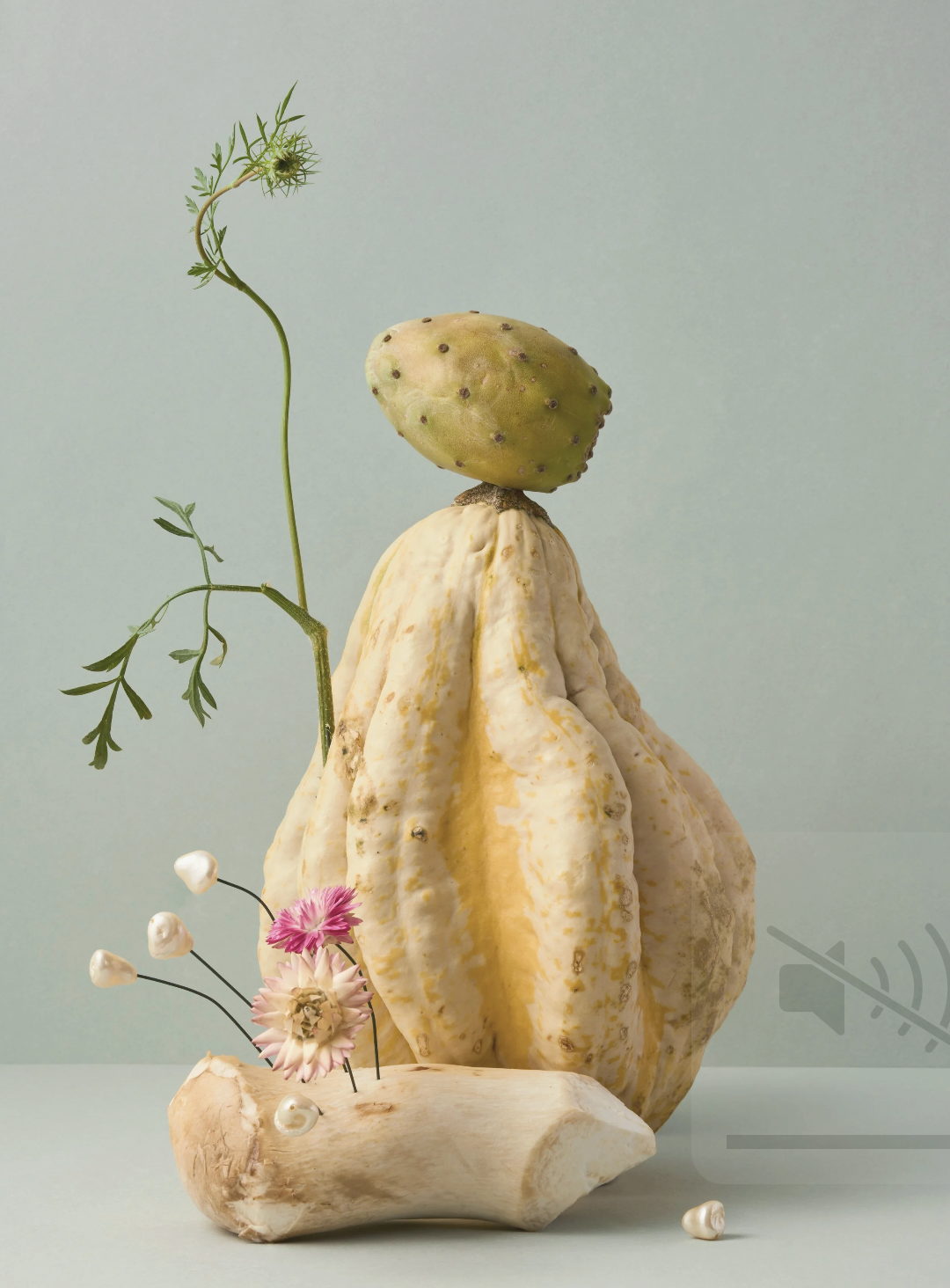 An art piece composed of a large yellowish gourd with a prickly pear cactus fruit on top, a mushroom base, a small pink flower, some white pearls, and green plant stems, against a plain light background.
