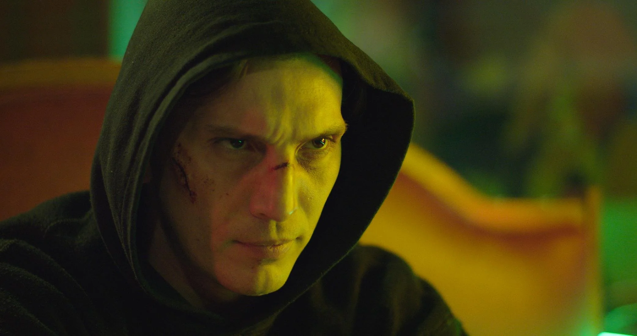 A young man with a serious facial expression, wearing a dark hooded sweatshirt, with street lighting in a colorful, blurred background.