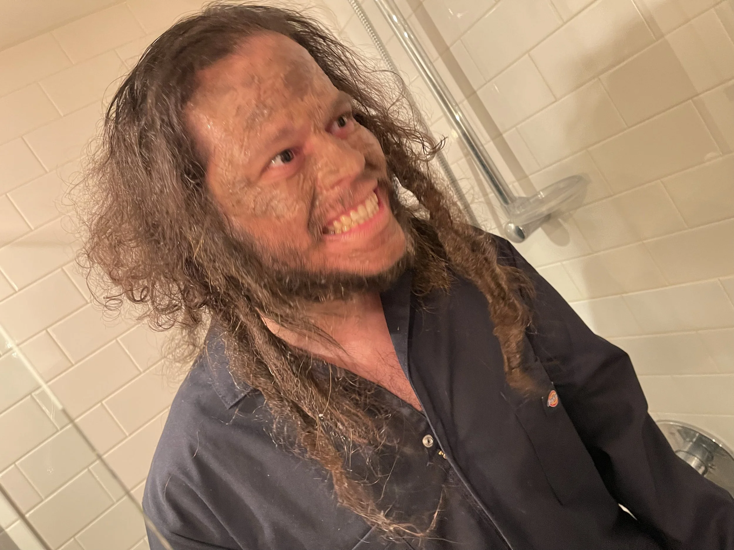 A man with long curly hair and a beard smiling in a bathroom with tiled walls, wearing a black uniform.