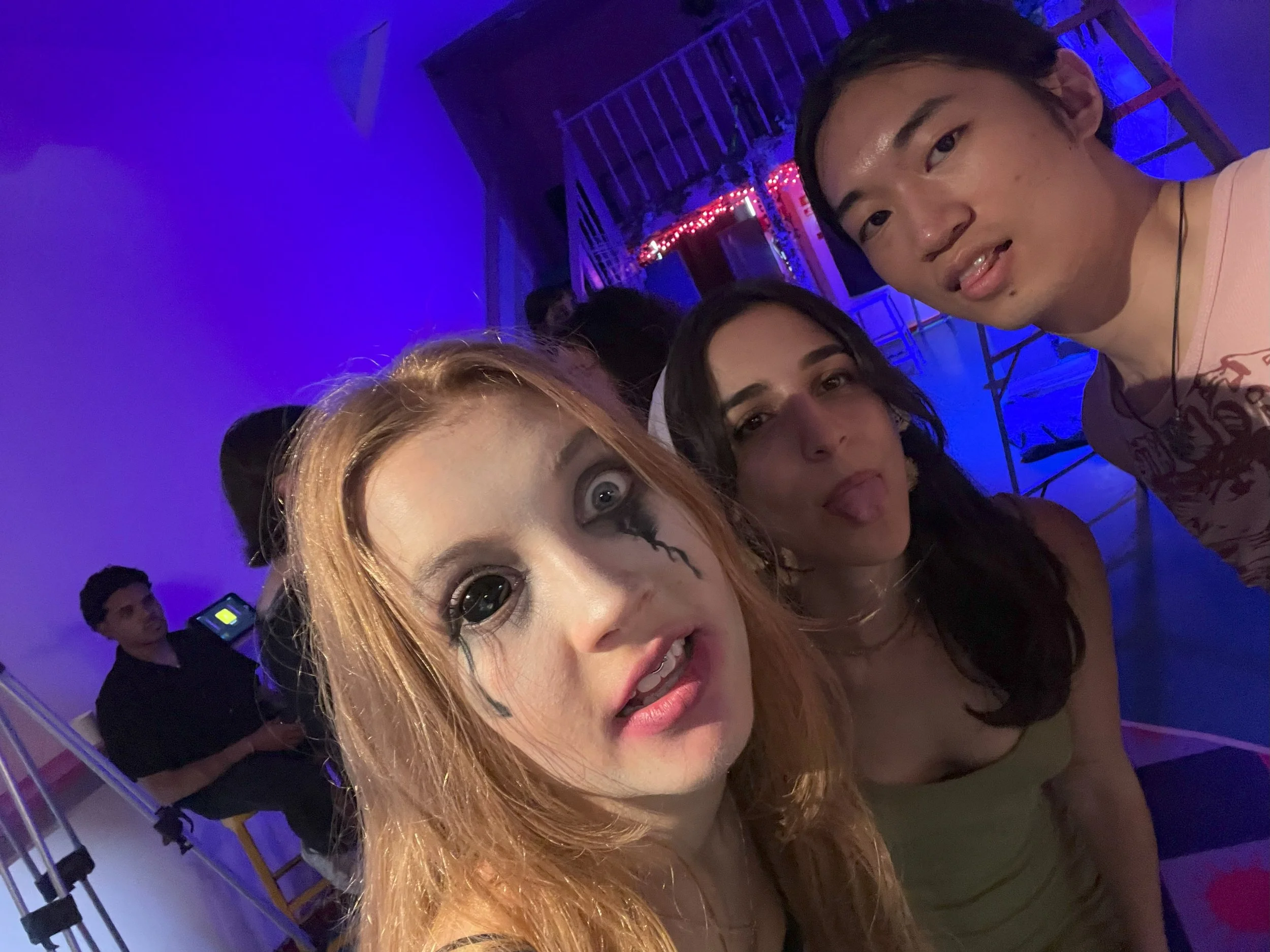A group of three young friends taking a selfie at a party or club with blue and red lighting. One person has orange hair and black makeup around their eyes, another has dark hair and is sticking out their tongue, and the third has dark hair and is making a surprised face.