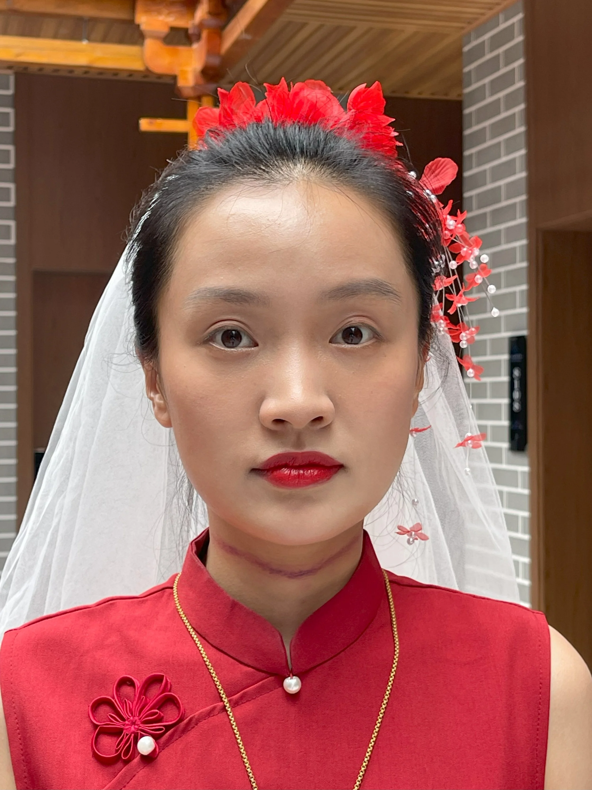 Woman wearing a red traditional dress with floral adornments and a veil, inside a room with wooden and brick interior.