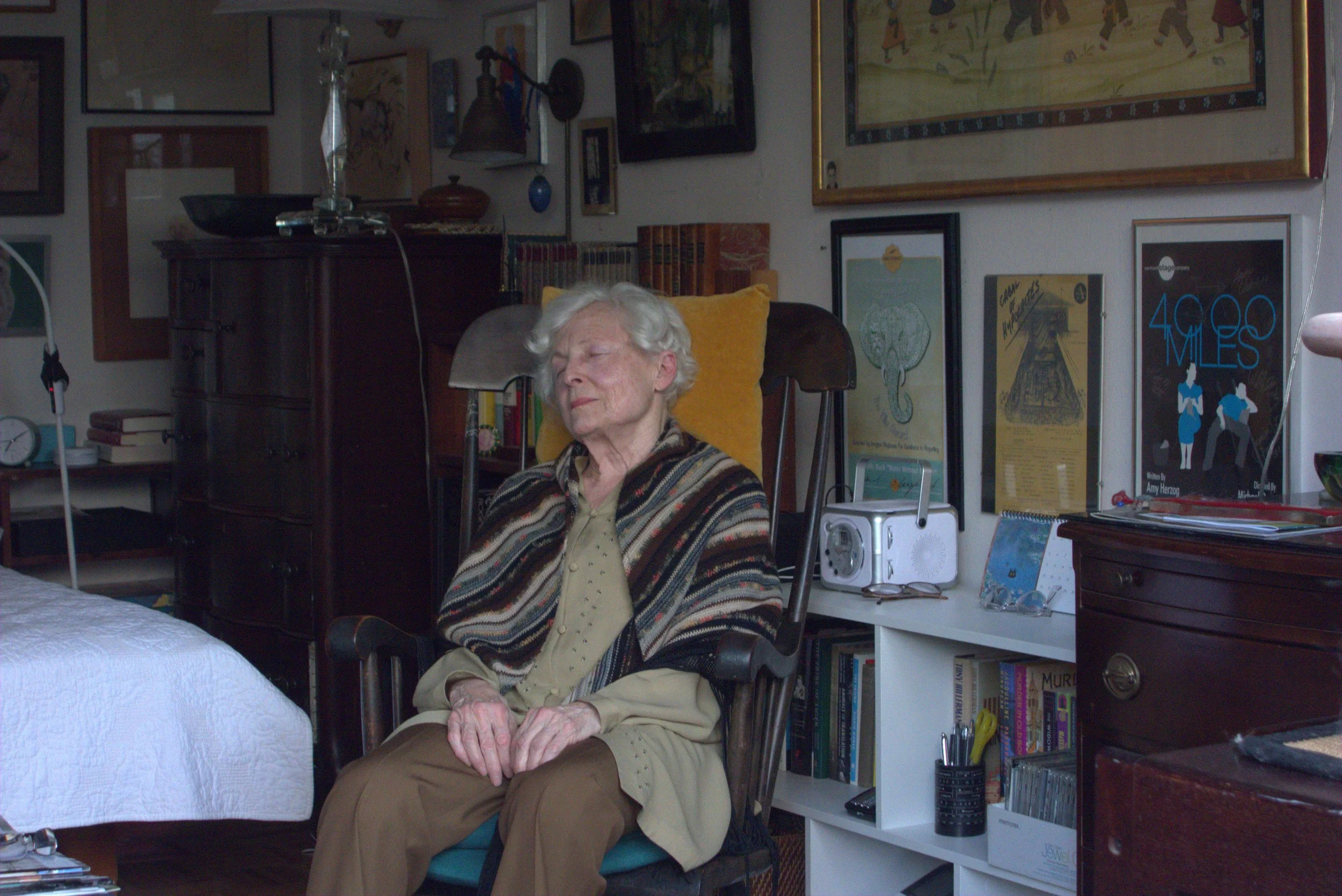 An elderly woman with white hair sitting in a room surrounded by framed artwork on the walls and books on shelves, with her eyes closed and hands resting on her lap.