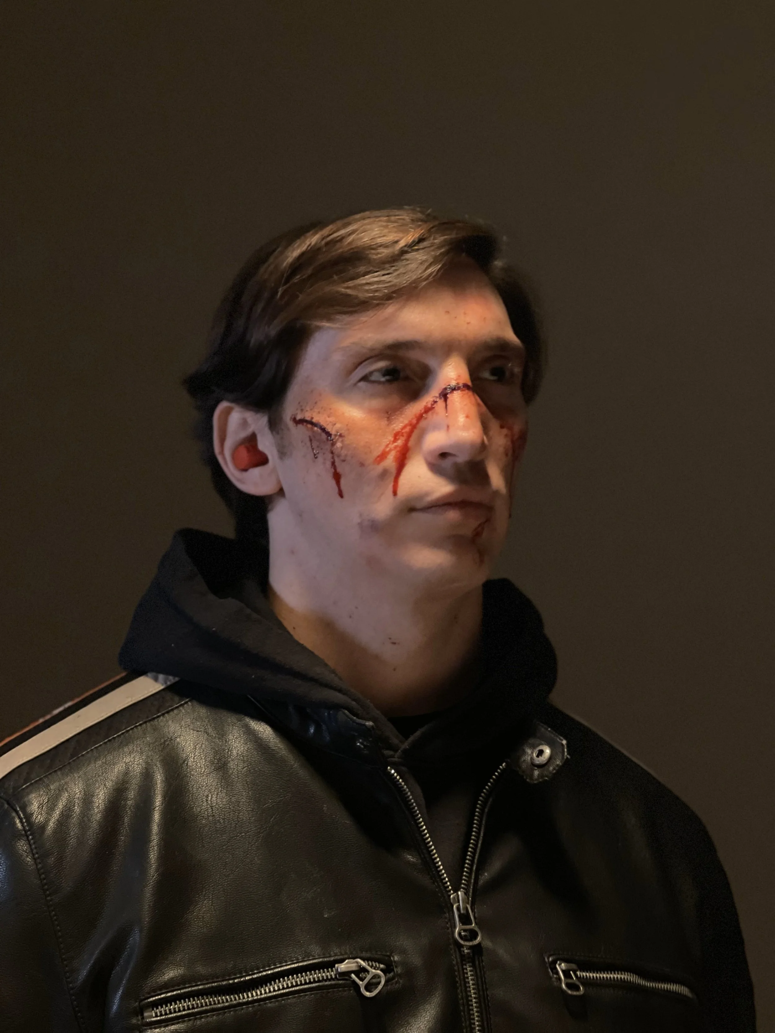 A person with short brown hair and light skin, wearing a black leather jacket with silver zippers, has pretend scars and blood on their face and neck, and is standing against a dark background.