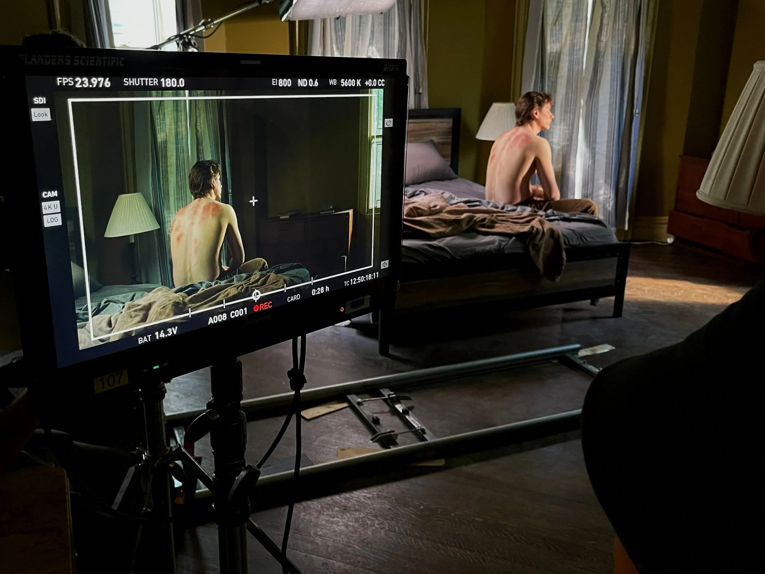 A film set capturing a scene of a shirtless middle-aged man sitting on a bed with a brown blanket, looking out of a window with sunlight streaming in, and a monitor displaying the scene in progress.
