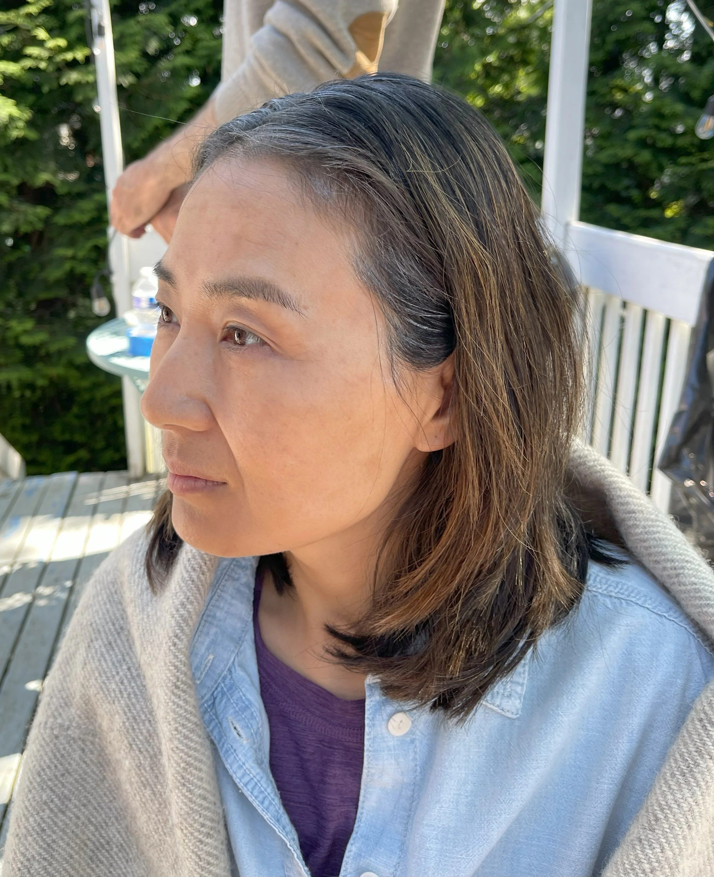 Close-up of a woman with shoulder-length brown hair, wearing a light blue denim shirt and a purple top, sitting outdoors with sunlight on her face and a green leafy background.