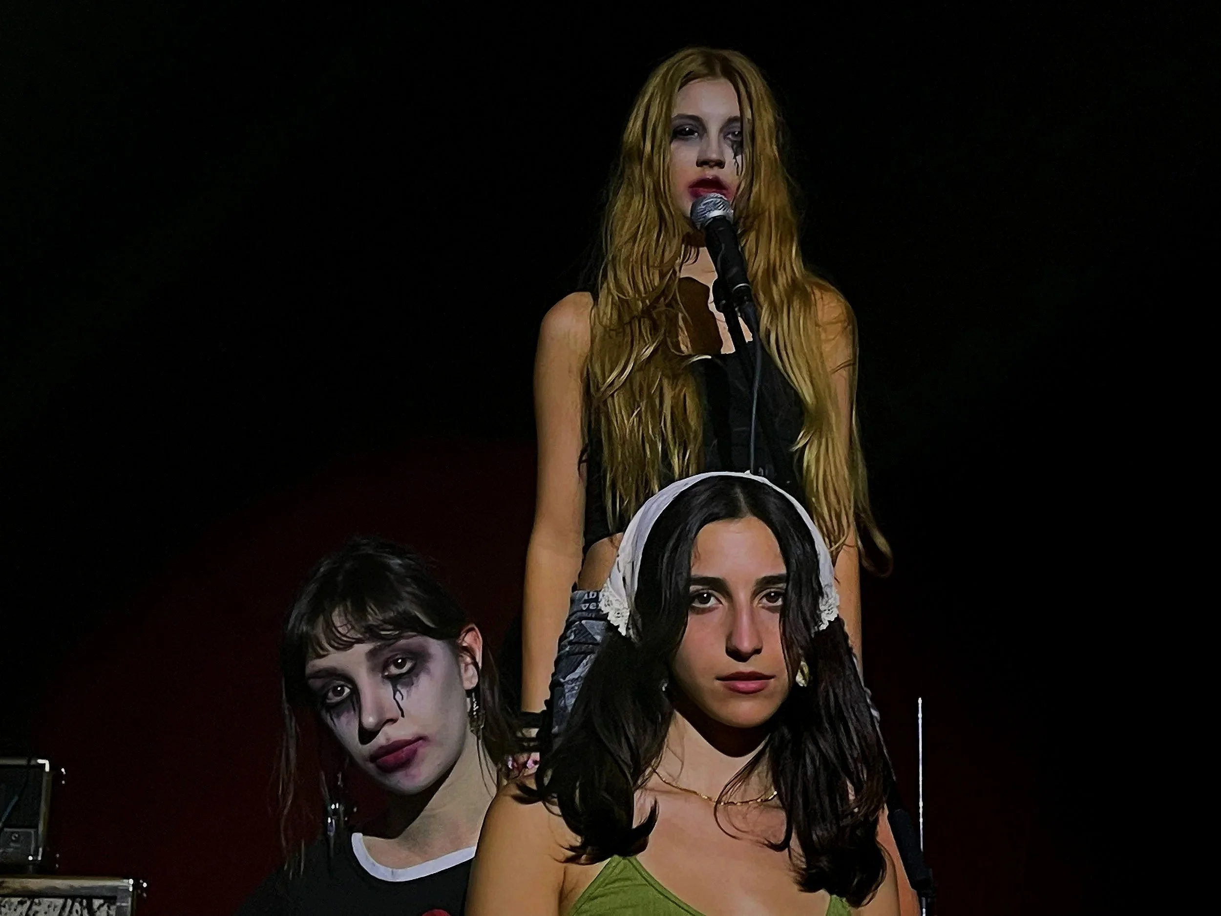 A woman with long blonde hair and dark eye makeup stands behind a microphone, with her face painted like a clown or mime, featuring dark lips and tears around her eyes. She is partially obscured by two other women in front of her, who have dark hair and dramatic makeup.