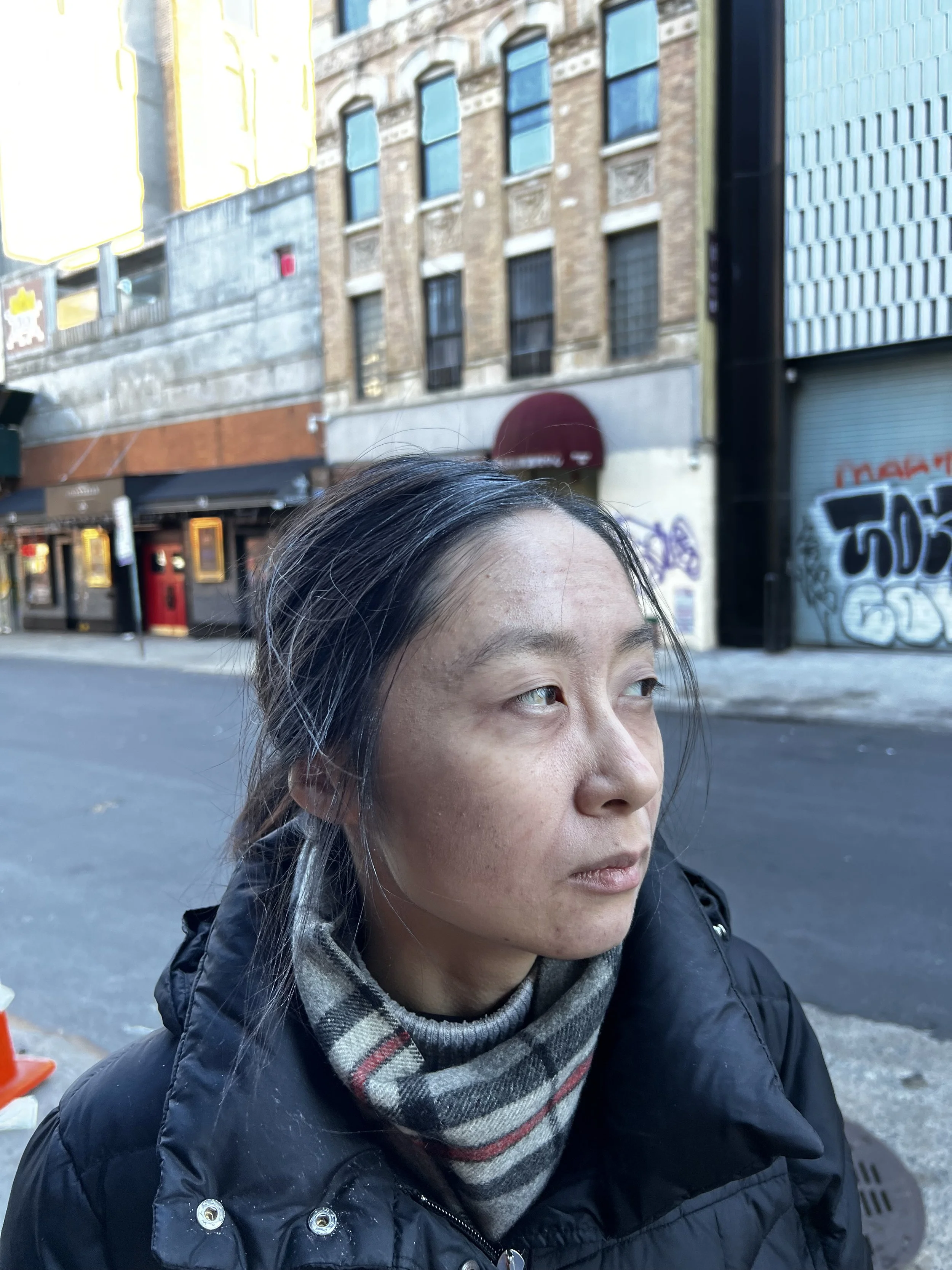 A woman with dark hair, wearing a black coat and a plaid scarf, looking to the right on a city street with old brick buildings and graffiti on a metal shutter in the background.