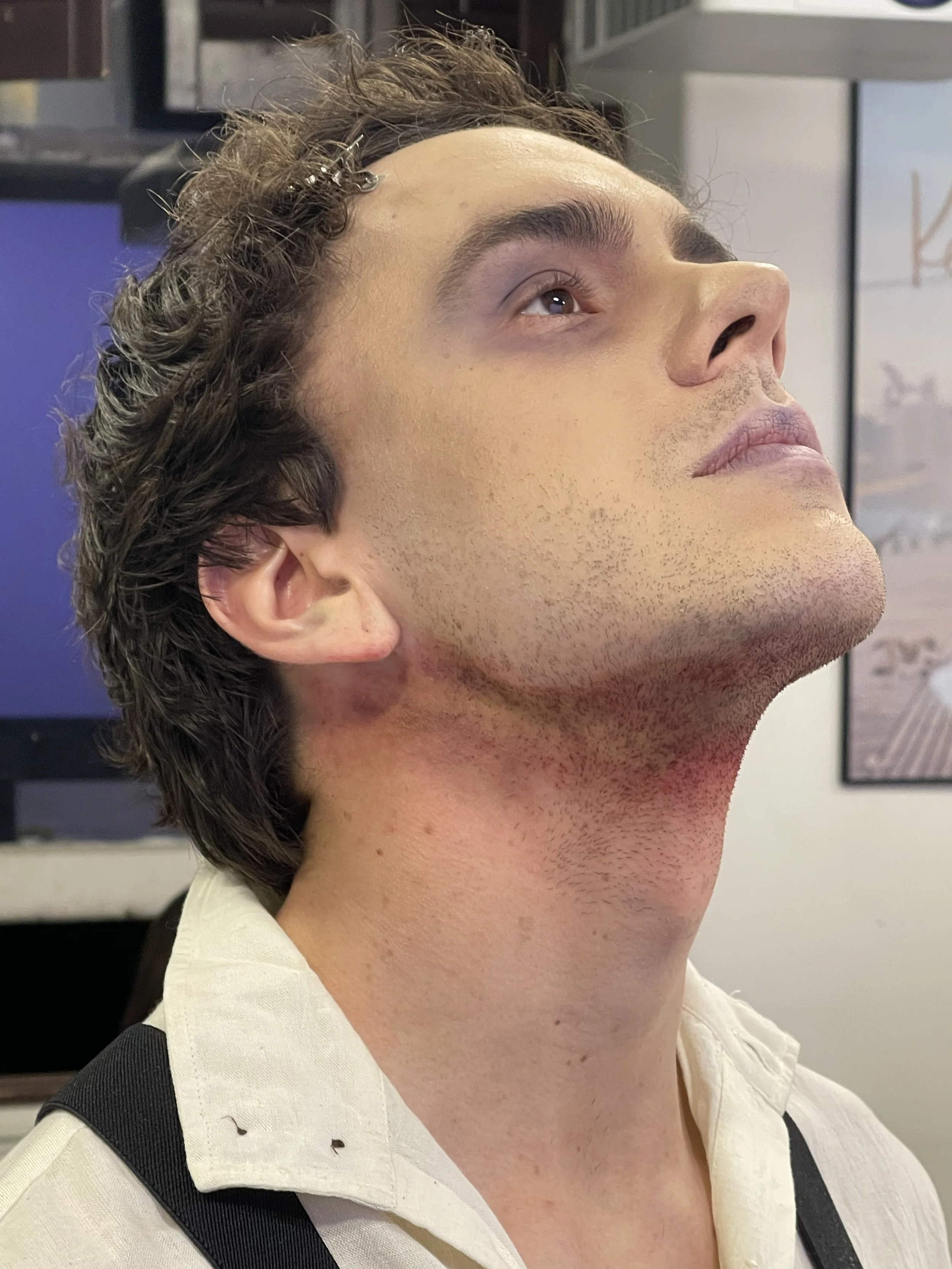 Close-up of a young man's face in profile, with short, curly brown hair, light skin, and a clean-shaven neck, wearing a white and black shirt, in an indoor setting with bookshelves and framed pictures in the background.