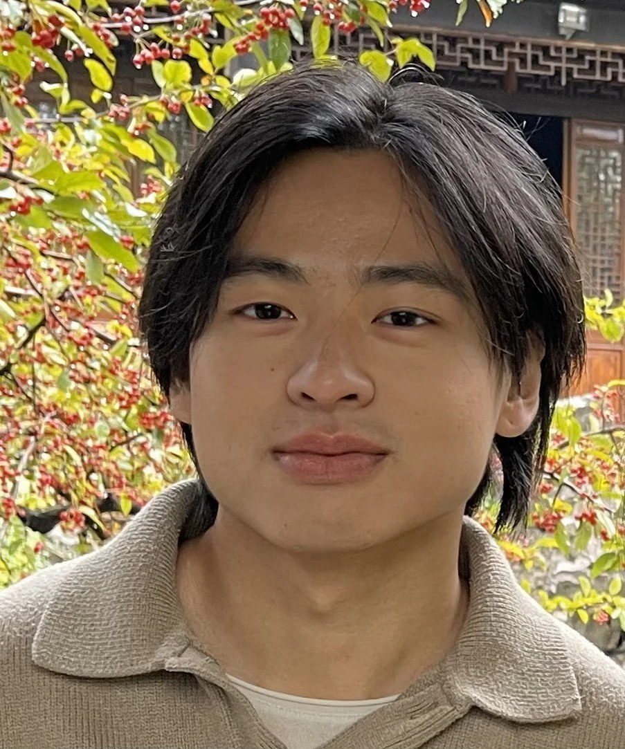 A young person with medium-length black hair standing outdoors near a bush with red and green leaves, with traditional Asian architectural elements in the background.