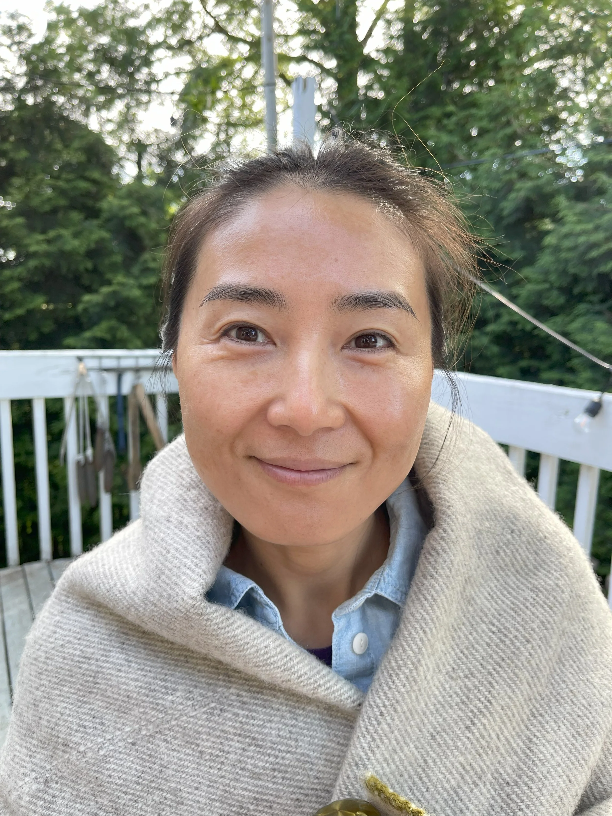 Close-up of a woman with dark hair and brown eyes smiling outdoors, wearing a beige sweater and a denim shirt, with a white fence and green trees in the background.