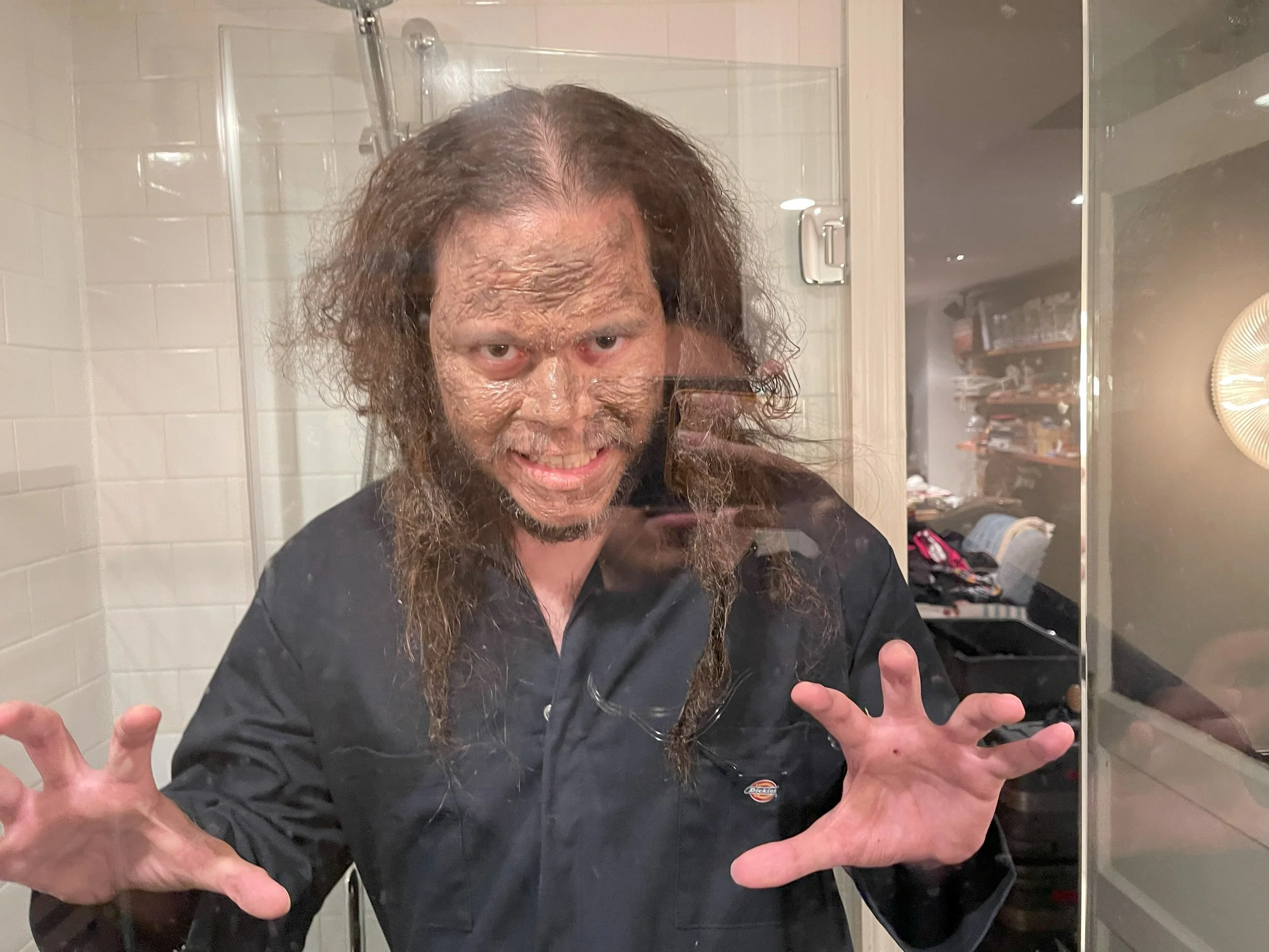 A man with long curly hair and a beard taking a selfie in front of a mirror in a bathroom. He is wearing a black shirt with a small logo on the chest, smiling, and making a playful gesture with his hands. The bathroom has beige tile walls, a glass shower door, and a shelf with toiletries in the background.