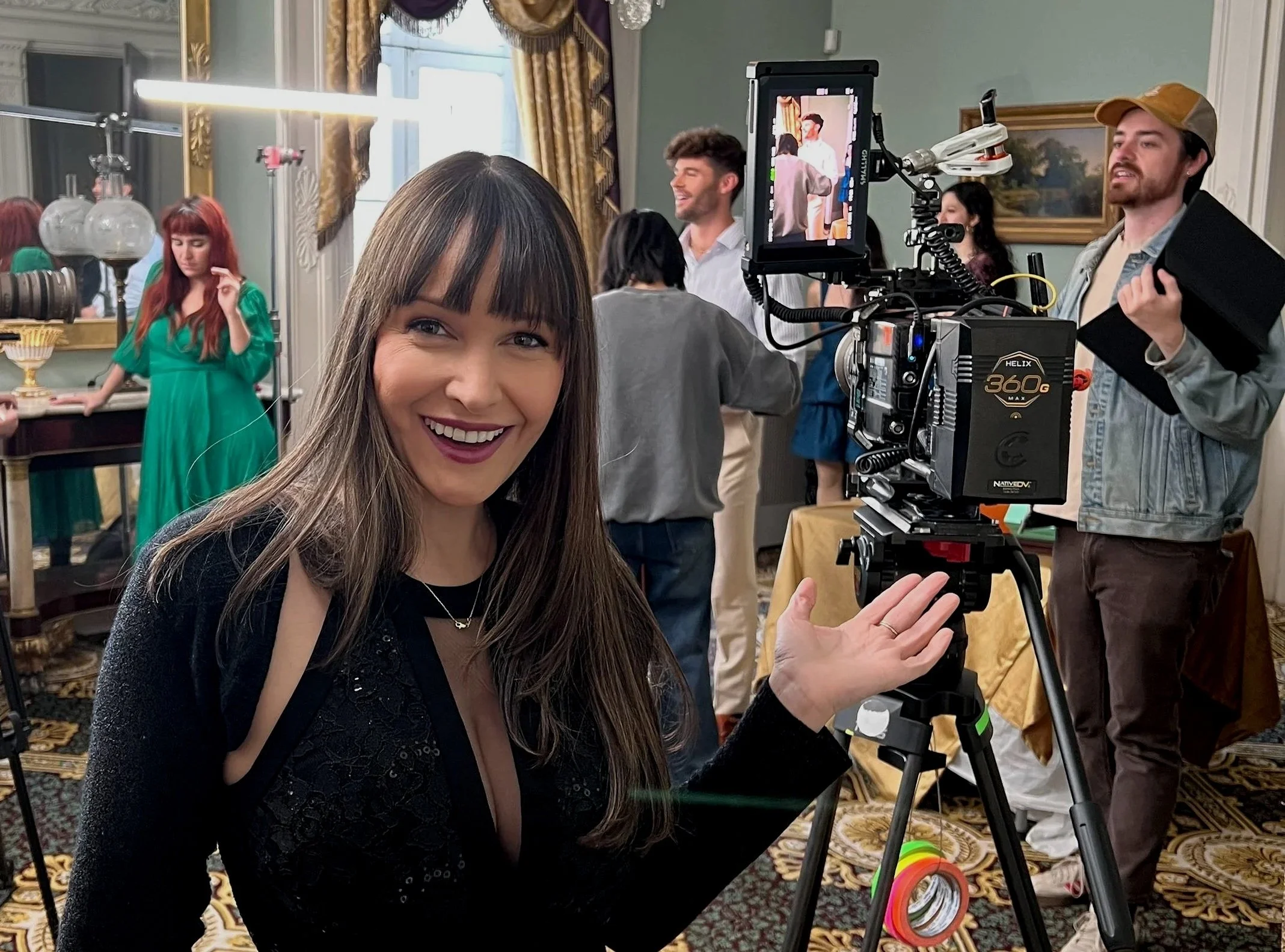A smiling woman with long brown hair and bangs takes a selfie in a room with ornate curtains and framed paintings, in front of professional filming equipment and crew members preparing to shoot a scene.