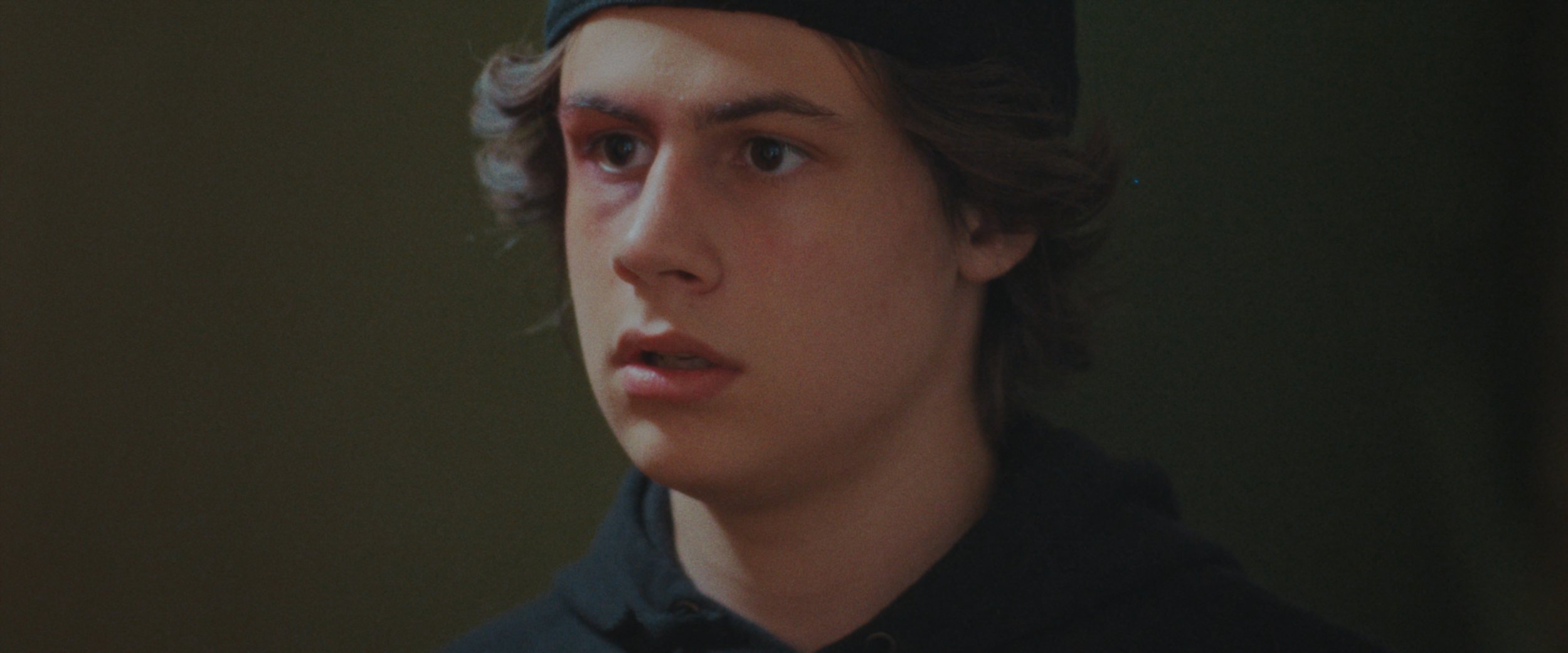 Close-up of a young man's face with curly brown hair, wearing a dark hoodie and a backward cap, looking to the side with a serious expression.