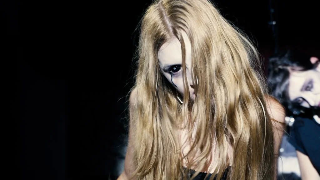 A woman with long, blonde hair, wearing dark makeup and a black top, looking downward in a dark setting.