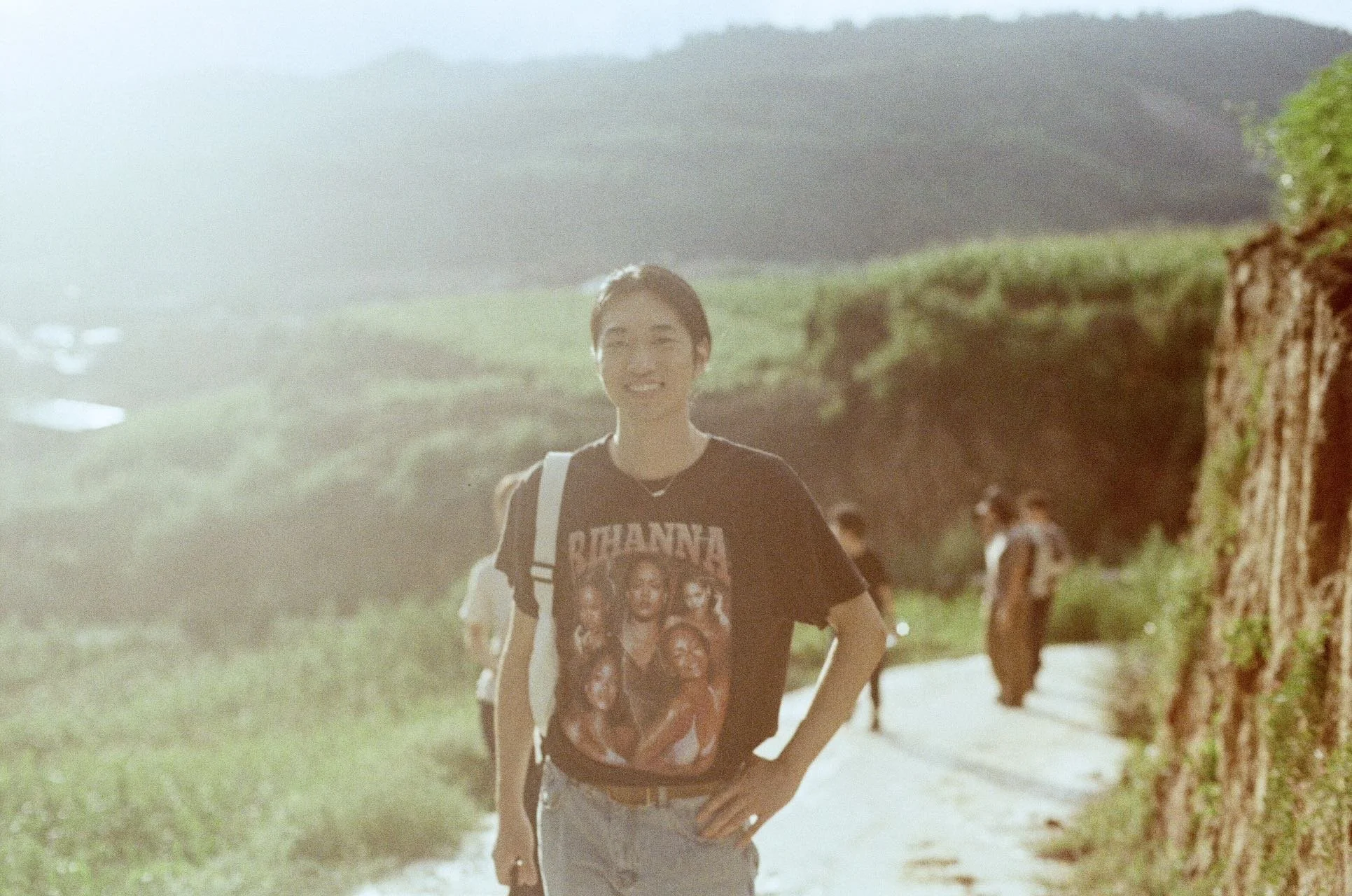 A young woman smiling on a sunny day walking outdoors, wearing a black T-shirt with a graphic and a backpack, with a scenic backdrop of green hills and other people in the background.