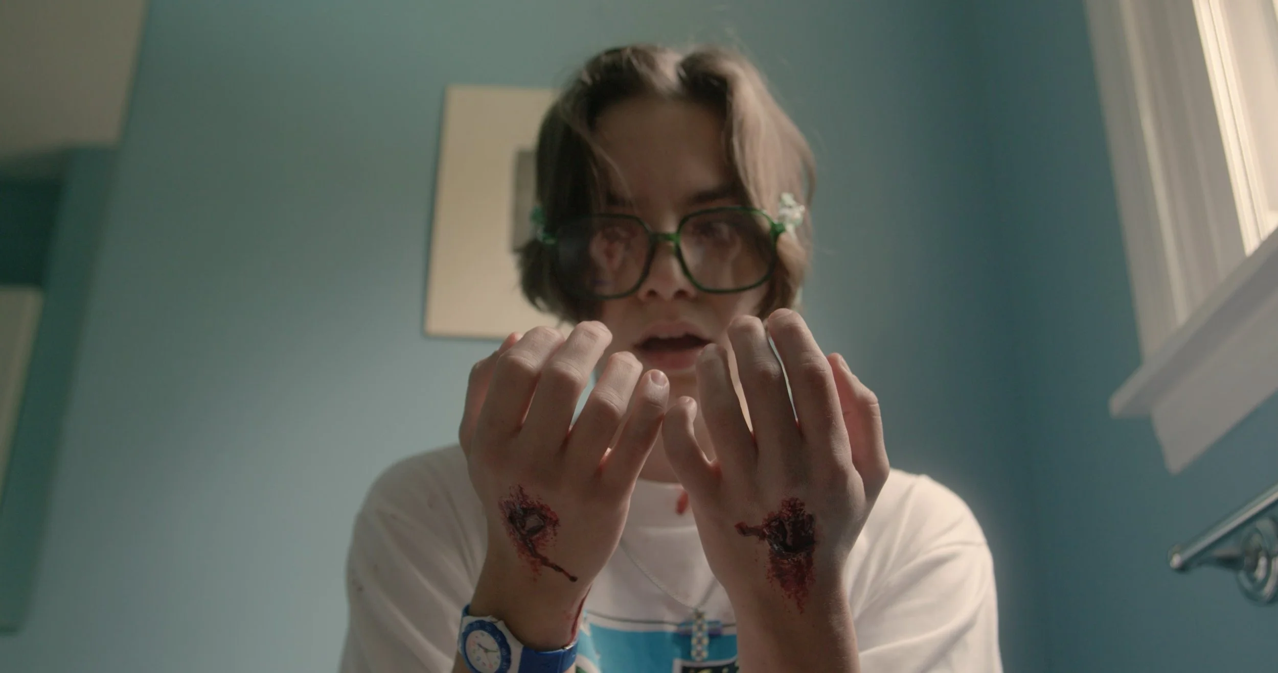 A young man with glasses and long hair looking down at his hands, which have blood on them, in a room with blue walls.
