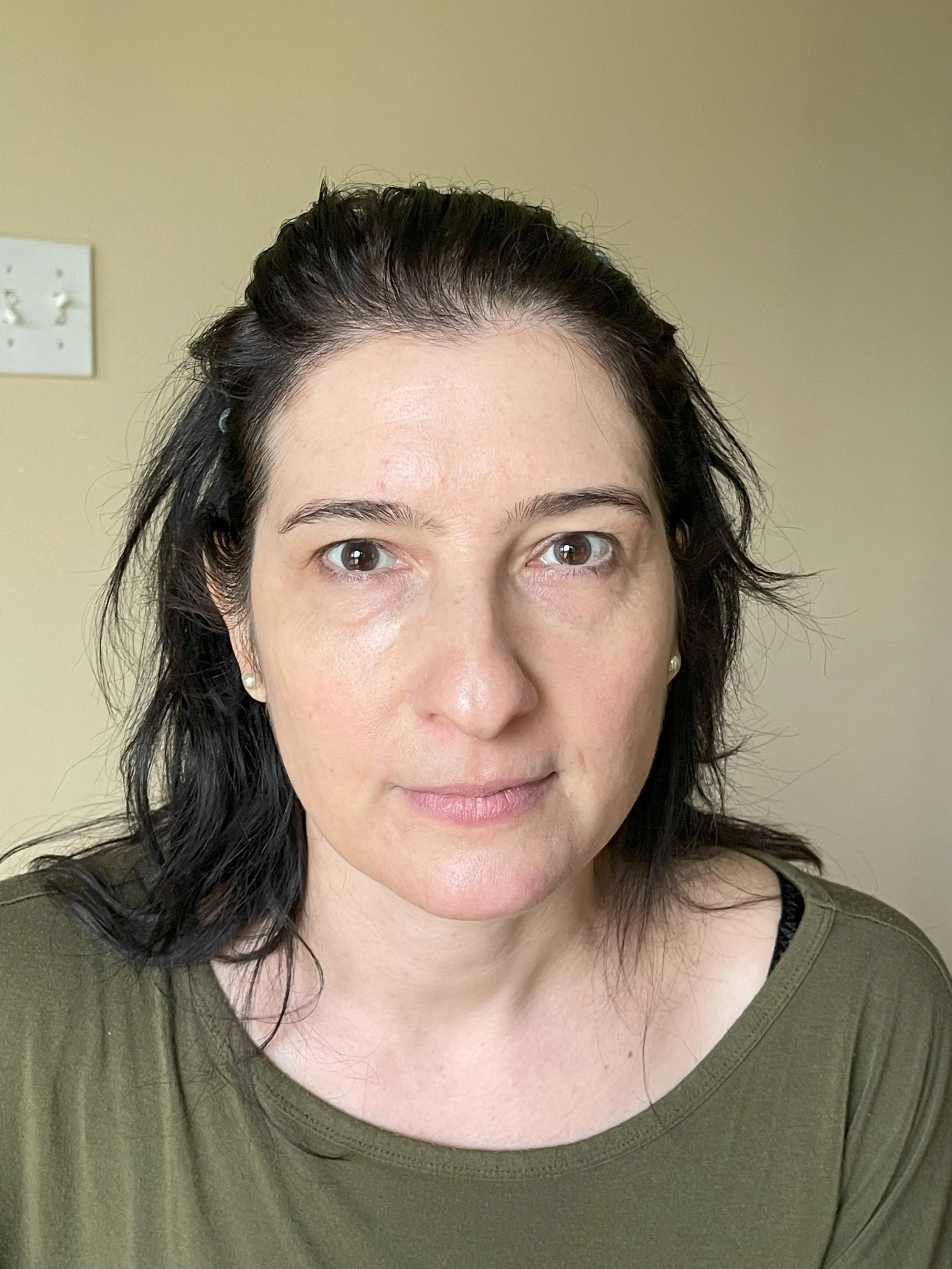 Close-up of a woman with dark, wavy hair, wearing a green top, in front of a plain beige wall with an electrical outlet visible in the background.