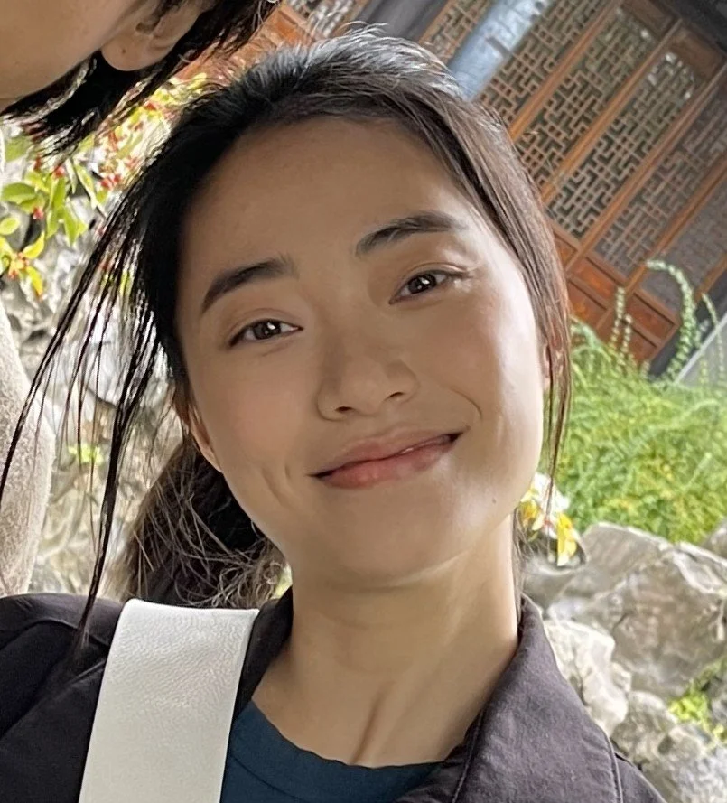 Young woman with dark hair smiling outdoors with rocks and plants in the background.