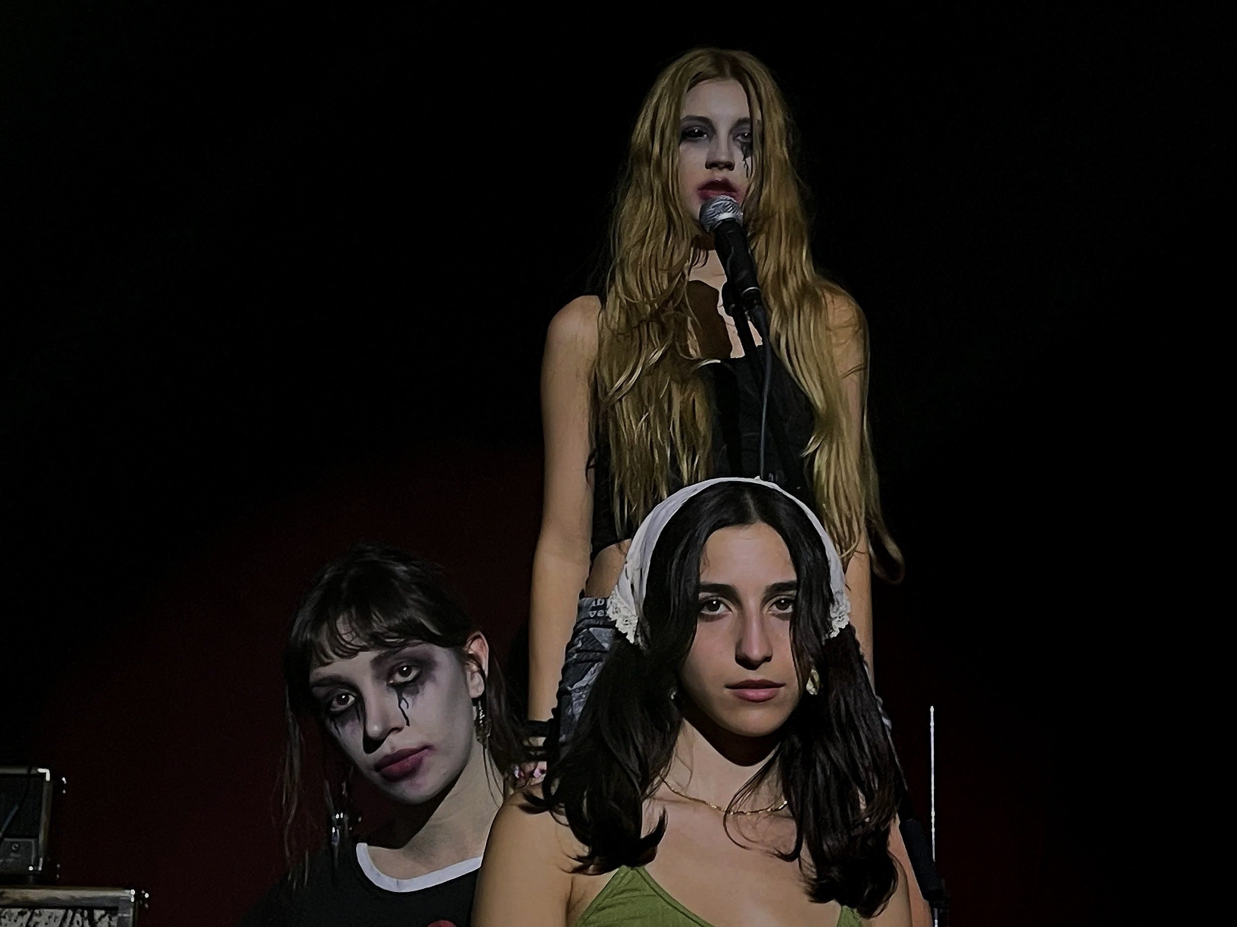 People dressed in avant-garde costumes with clown makeup, standing close together against a black background, in a theatrical or artistic setting.