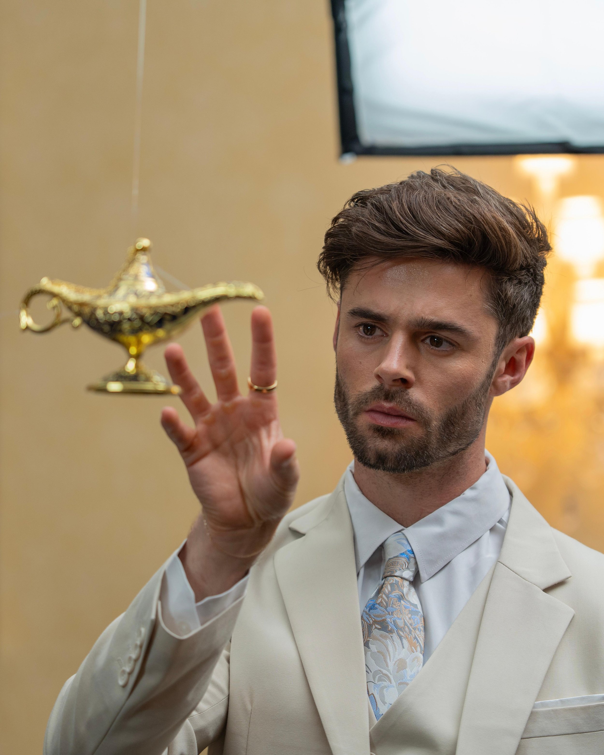 A young man in a cream-colored suit holding a small ornate gold lamp in his left hand.