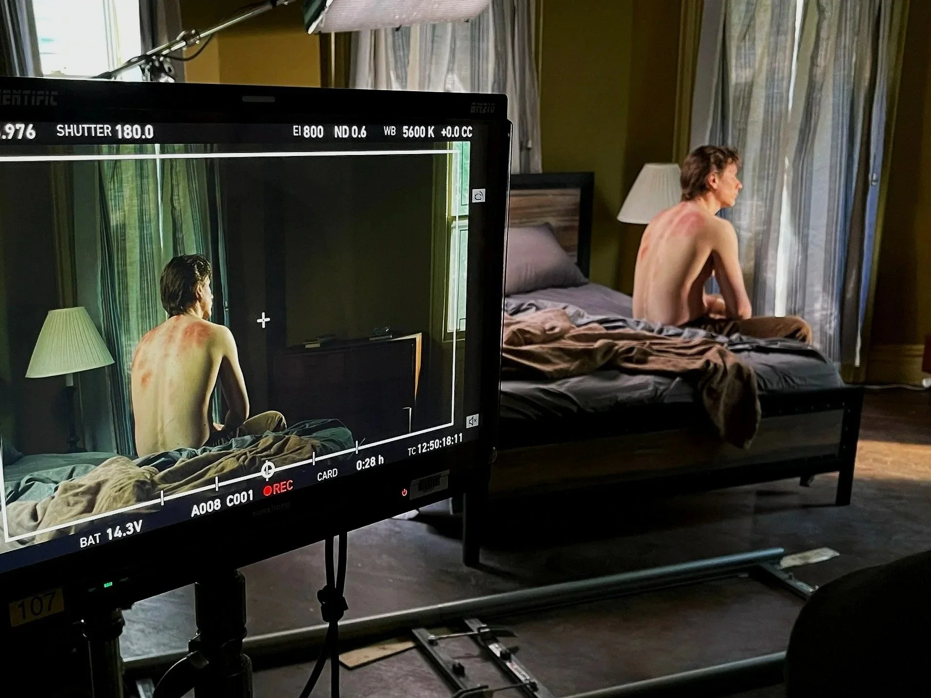 On a film set, a camera monitor shows a scene of a shirtless man sitting on a bed, with scars or bruises on his back, in a room with curtains and a bedside lamp.