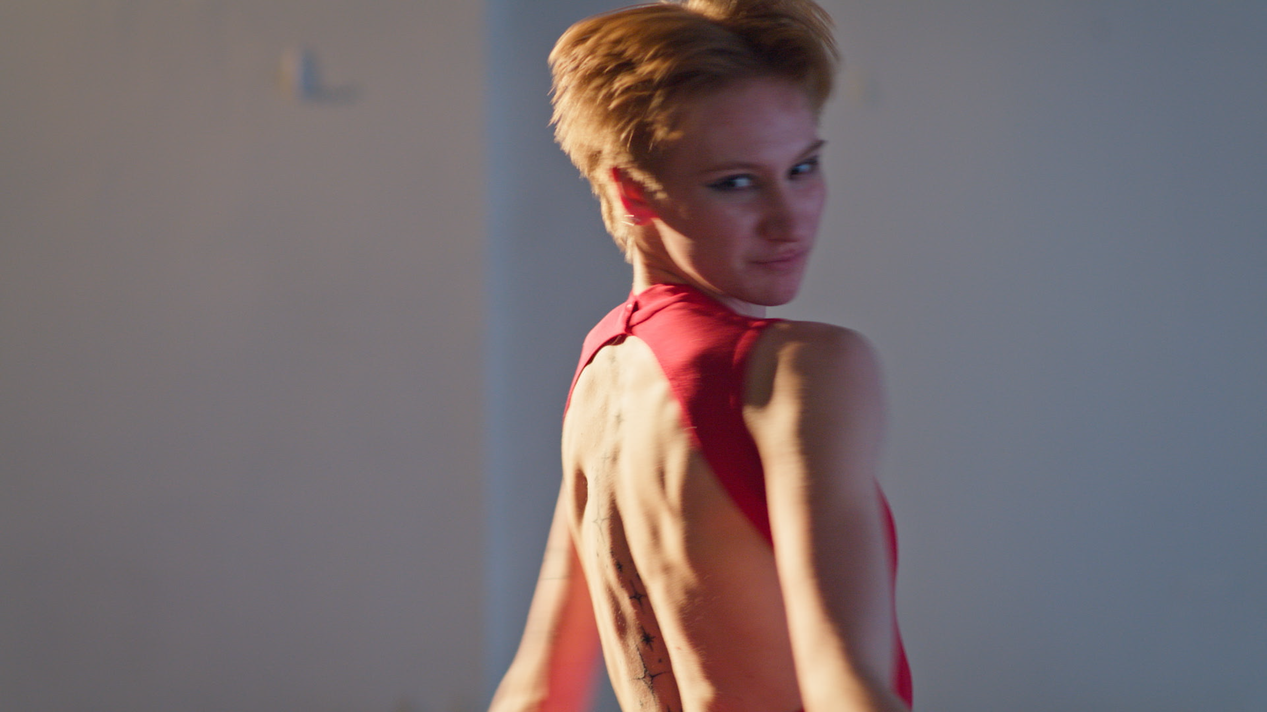 A woman with short blonde hair wearing a red top, looking over her shoulder at the camera, with a neutral background.