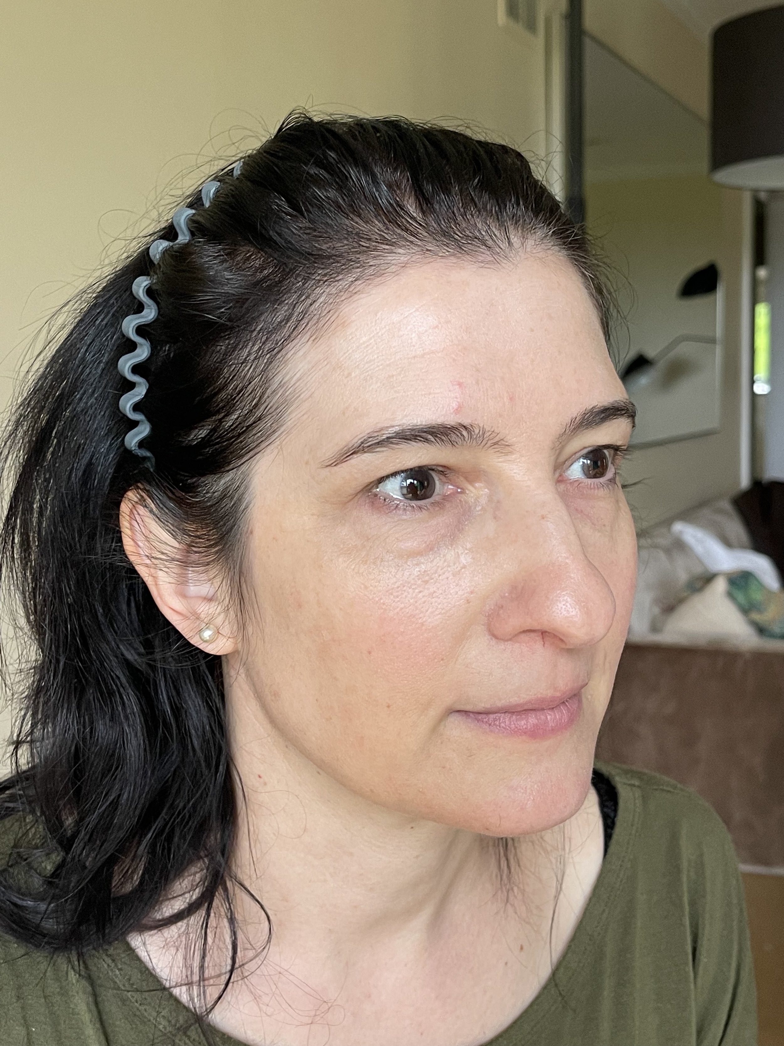 A close-up of a woman's face and hair, with her dark hair partially pinned back with a blue curl clip, and her fair skin visible. She is wearing a green top and a pearl earring.