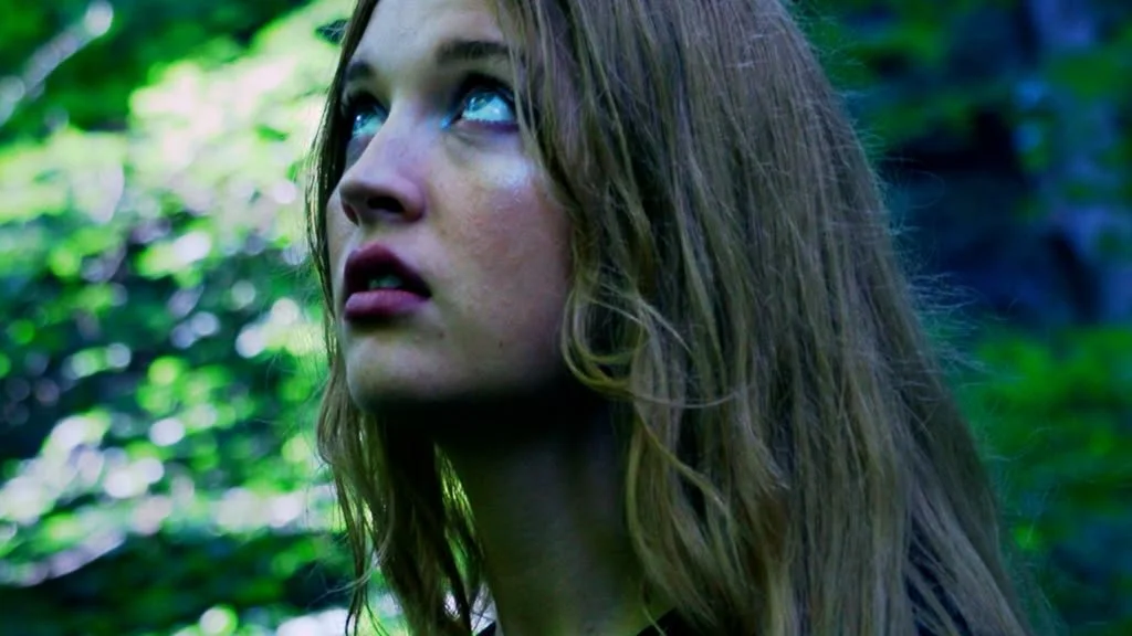 A woman with long brown hair looking upwards in a green outdoor setting.