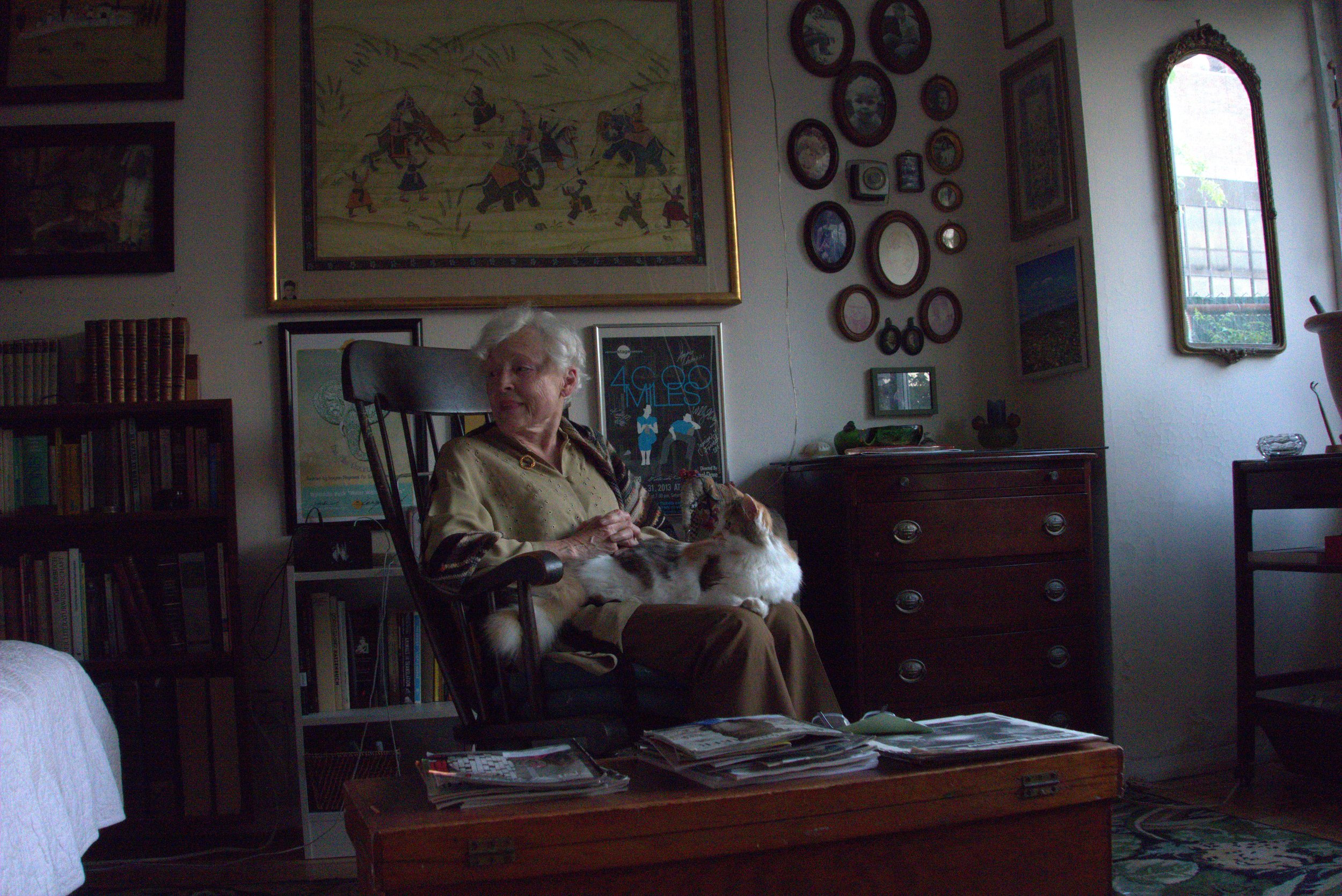 An elderly woman with white hair sitting in a chair, looking at a dog lying on her lap. The room has a dark wooden dresser, a bookshelf filled with books, and various framed pictures and artwork on the beige wall. Sunshine comes through a large arched window on the right side of the room.