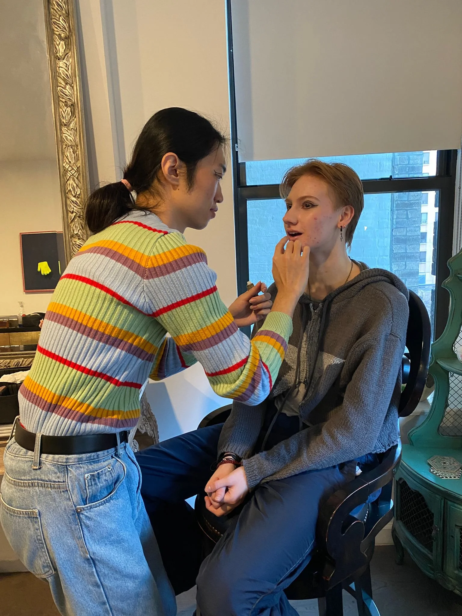 A person with dark hair in a ponytail is applying makeup to a seated person with short, light brown hair, in a room with a large window and a decorative mirror.