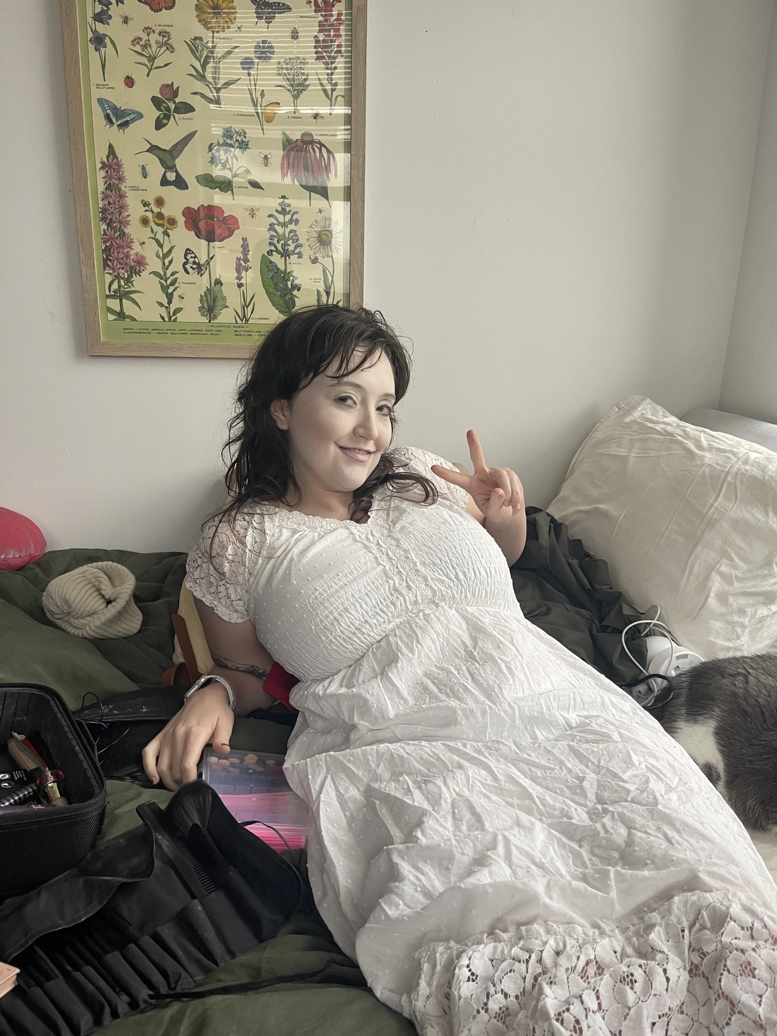 A woman lying on a bed, smiling and making a peace sign with her fingers, with various personal items and a floral poster on the wall behind her.