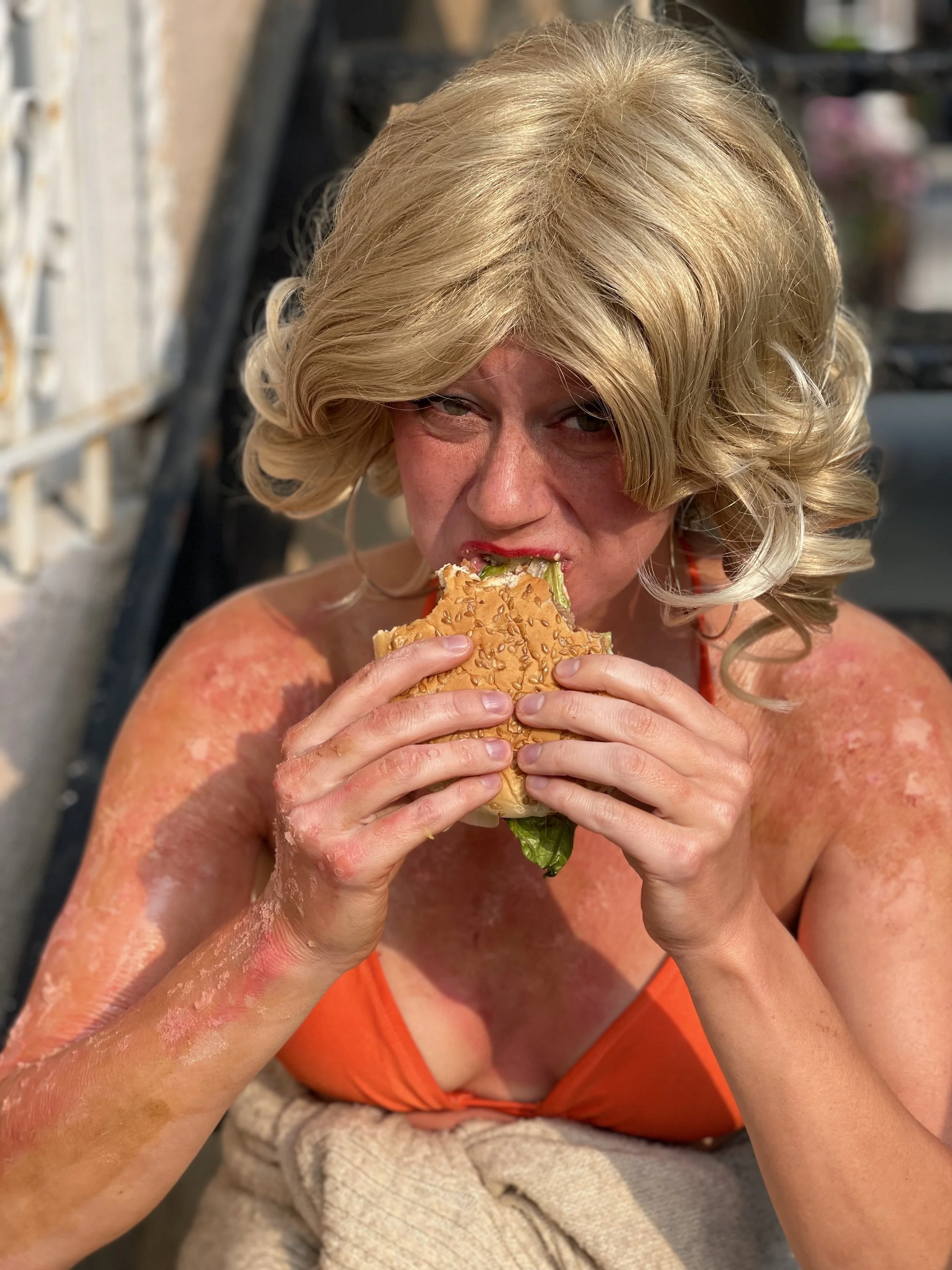 A woman with curly blonde hair and red lipstick eating a sesame seed bun with lettuce and tomato.