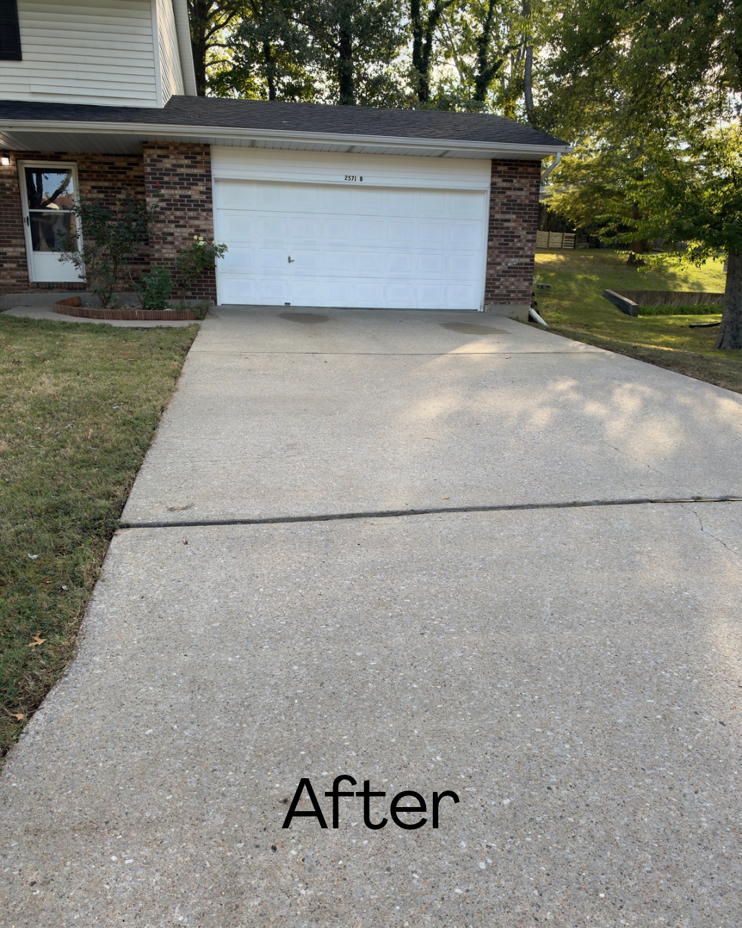 Clean, newly sealed concrete driveway in front of a brick house with a garage door, a small garden with plants, and trees in the background.