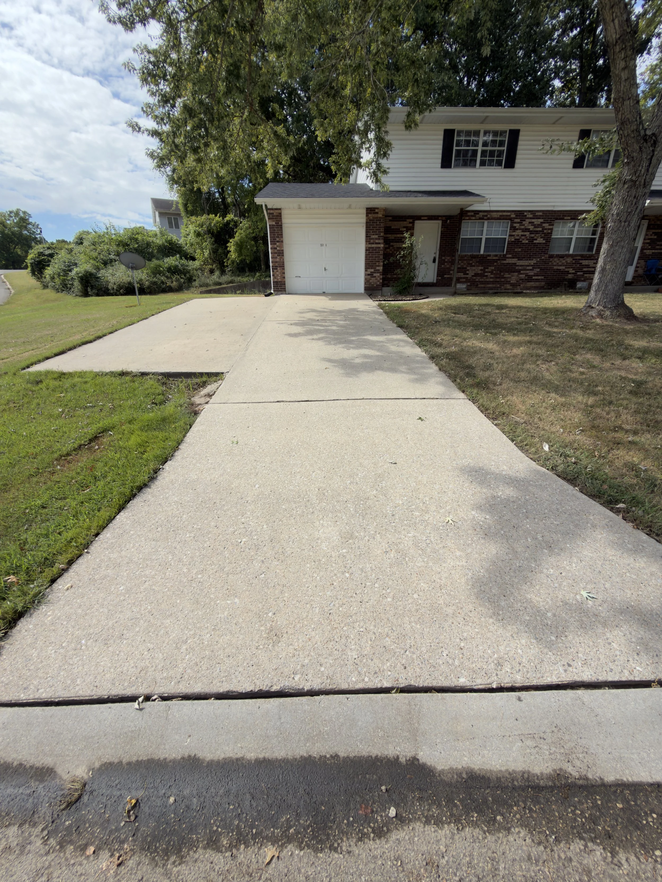 Photo of a sidewalk leading to the entrance of a house with a brick and white siding exterior, a white garage door, and a front door. There is a tree with green leaves on the right side and a driveway on the left. The sky is partly cloudy.
