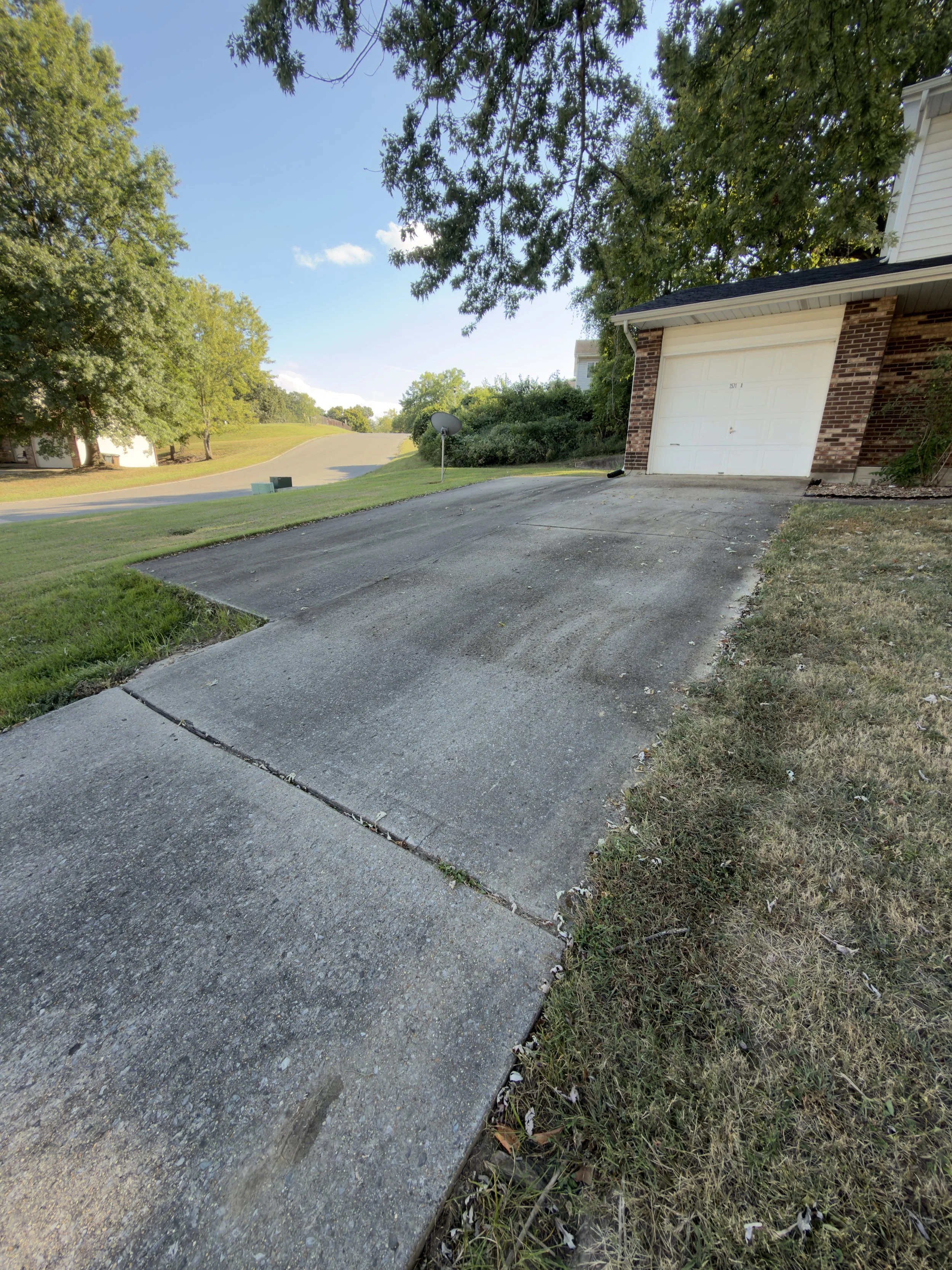 The image shows a residential driveway leading up a hill, located next to a brick house with an attached garage. There are trees on either side of the driveway, providing shade. The sky is mostly clear with a few small clouds.