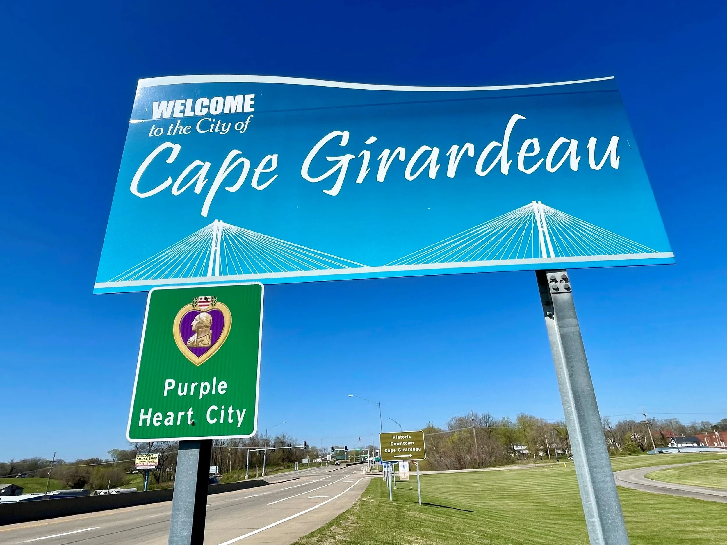 Blue highway sign welcoming to Cape Girardeau, Missouri, with a graphic of a bridge and a smaller green sign indicating the city is in the Heart of the Midwest.