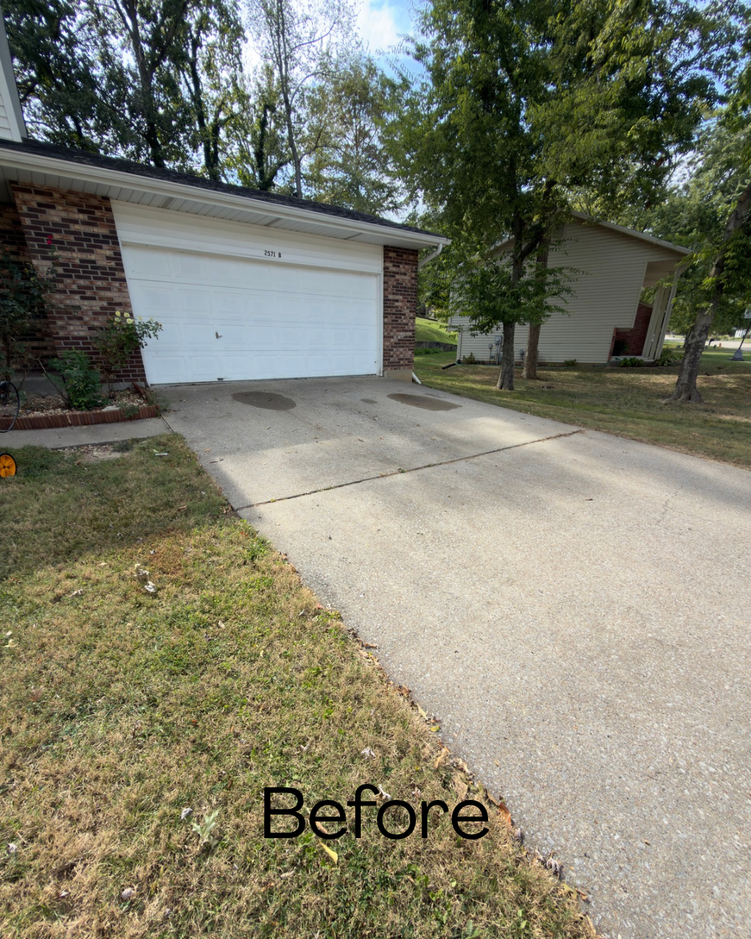 A concrete driveway leading up to a white garage door with a brick border on one side, a small landscaped area with plants, and trees in a suburban neighborhood, with the word "Before" in black text at the bottom.