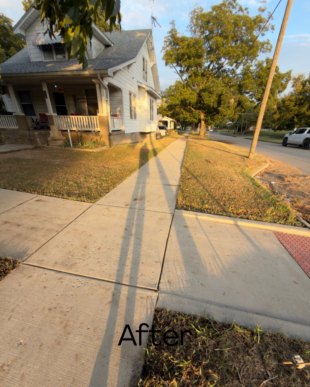 Sidewalk and yard with shadows cast by a utility pole, with a white house and cars parked on the street, taken in the late afternoon or early evening.