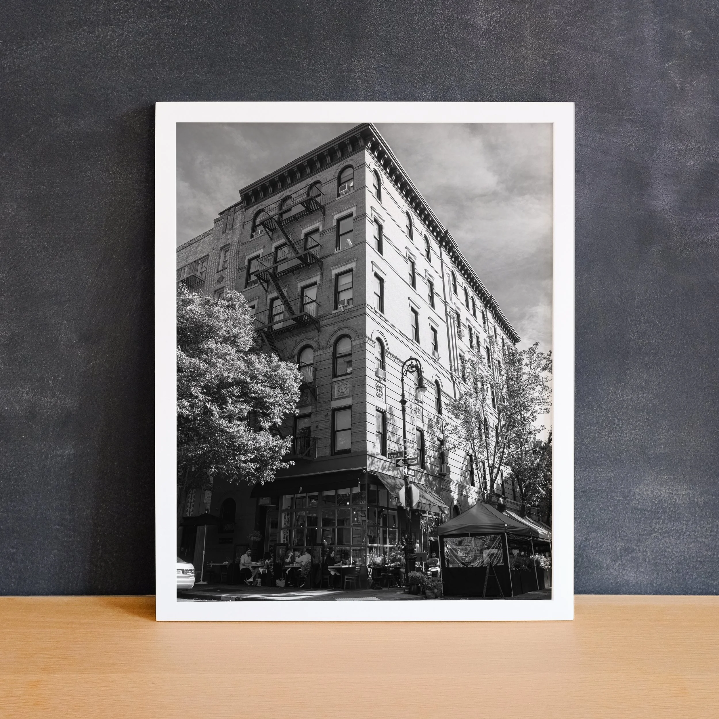 White framed Bedford and Grove fine art print leaning against a dark charcoal wall on a light wood surface.