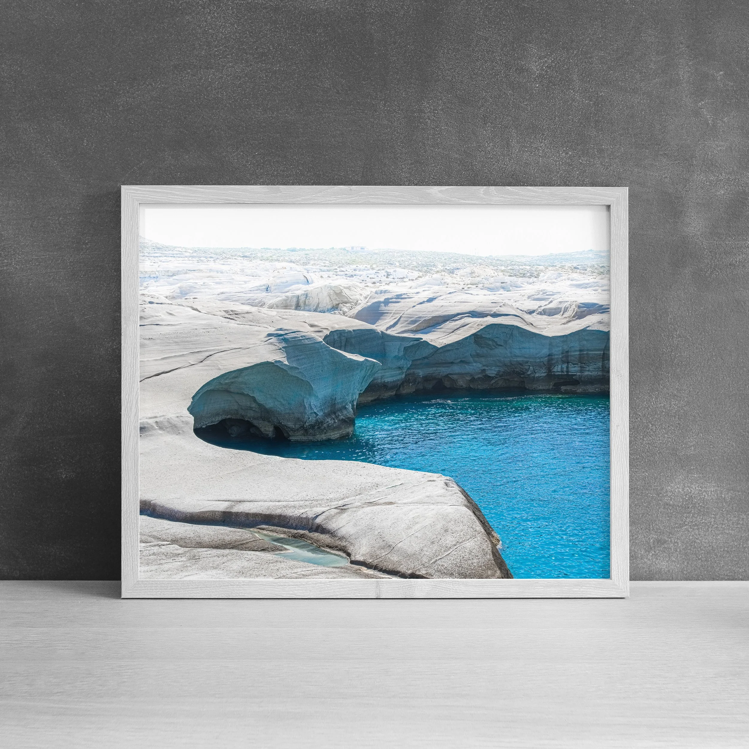 White framed landscape photography print of the iconic white cliffs and blue waters of Sarakiniko Beach in Milos, leaning against a dark charcoal wall.