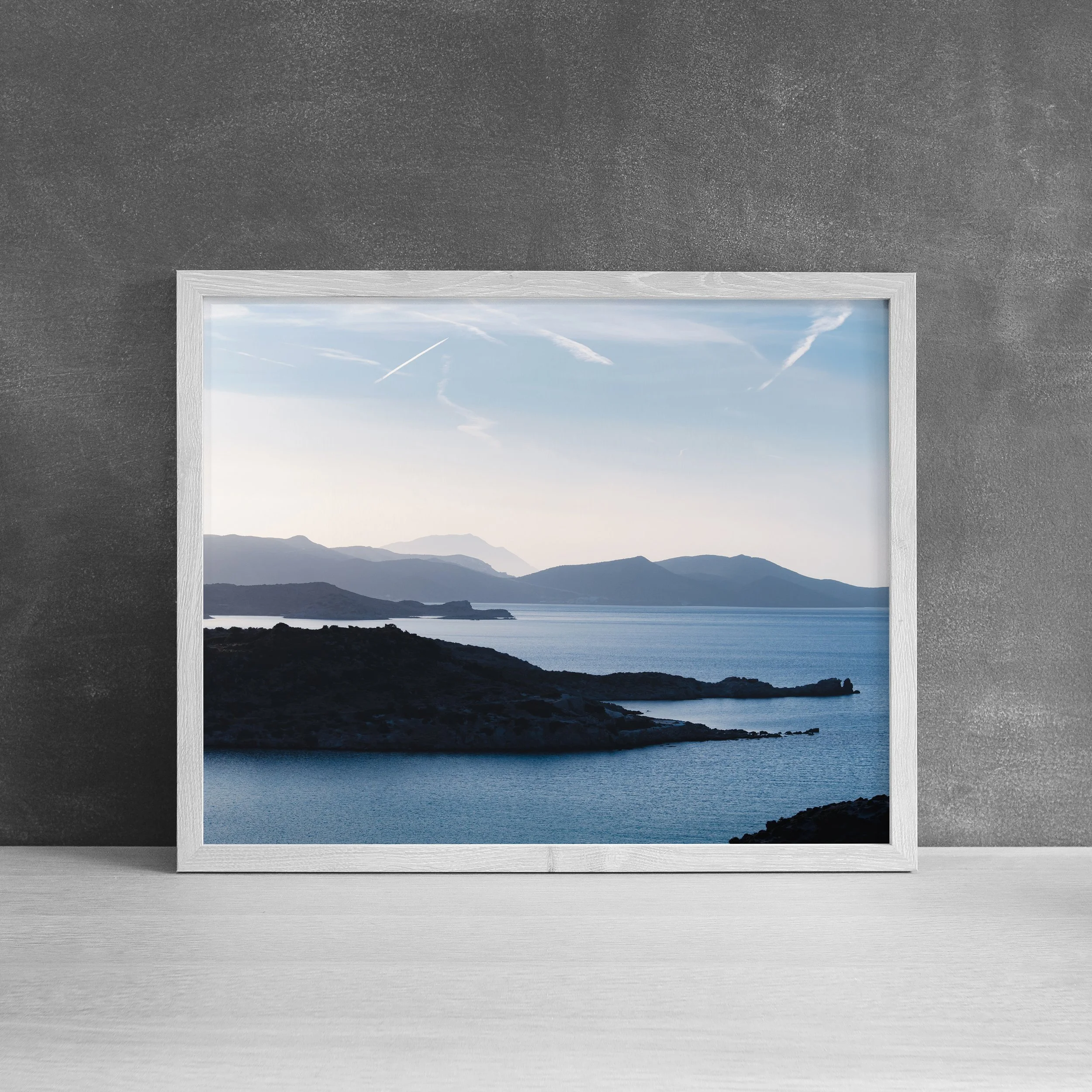 Framed fine art print of Milos, Greece coastal mountains and blue Aegean Sea displayed against a textured grey wall.