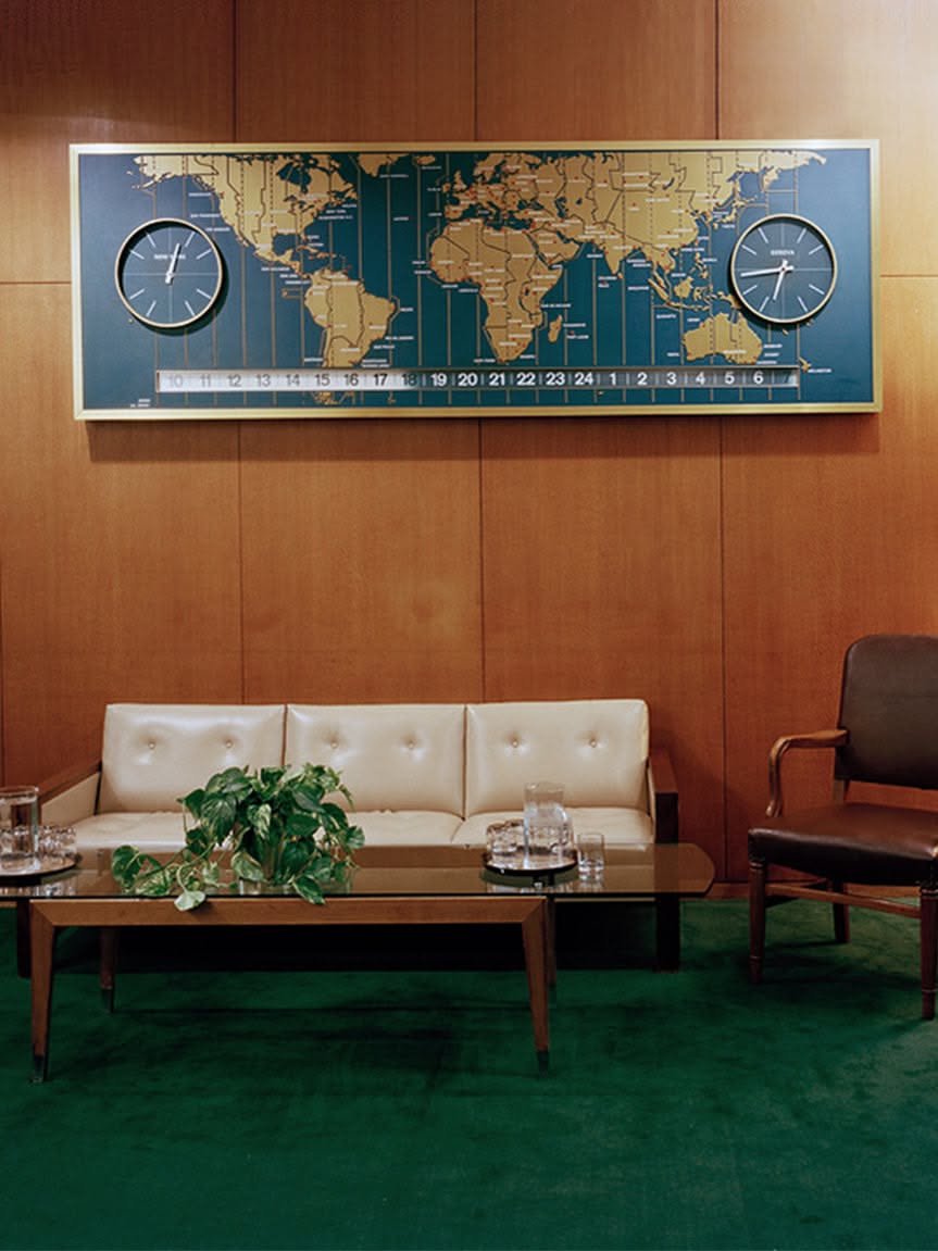 The existing brand’s wood-paneled office scene, with cream seating and green carpet.