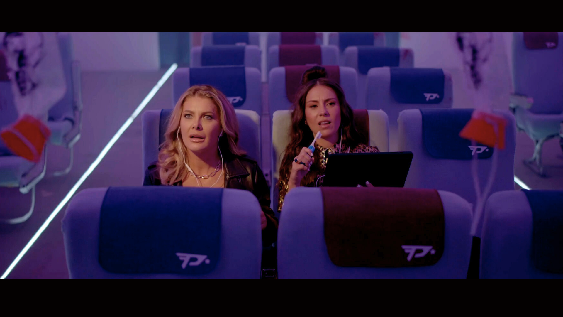 Two women sitting in an airplane cabin looking surprised and confused, with blinking interior lights and bags hanging on the side.