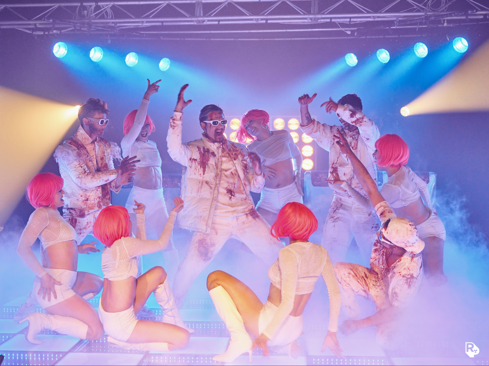 Group of performers on stage with colorful lights, some wearing pink wigs, white outfits, and sunglasses, dancing and posing dynamically.
