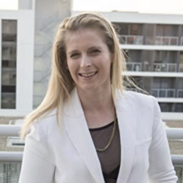A woman with long blonde hair smiling outdoors on a balcony, wearing a white blazer and a black top with a gold chain necklace, with a cityscape in the background.