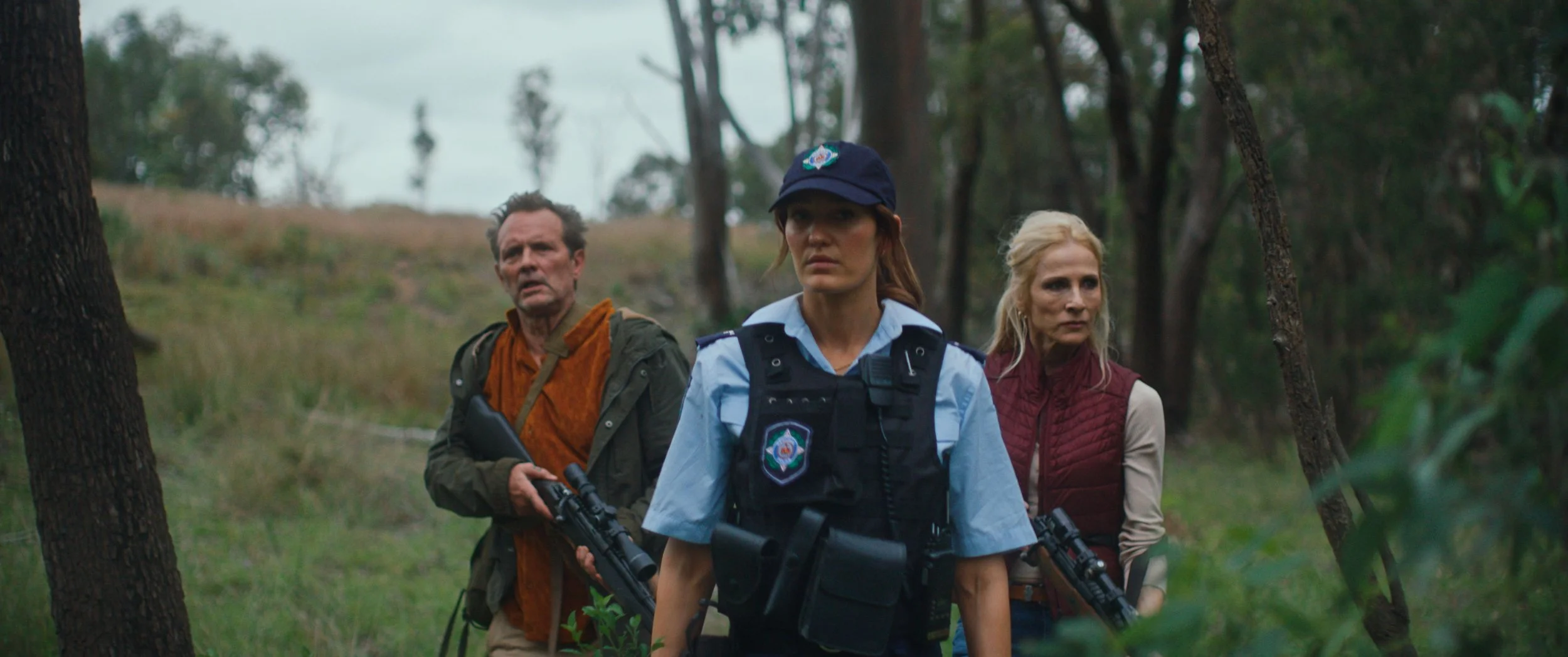 Three people walking through a wooded area, with the central figure being a woman in police uniform holding a rifle, flanked by a man and a woman in civilian clothing.